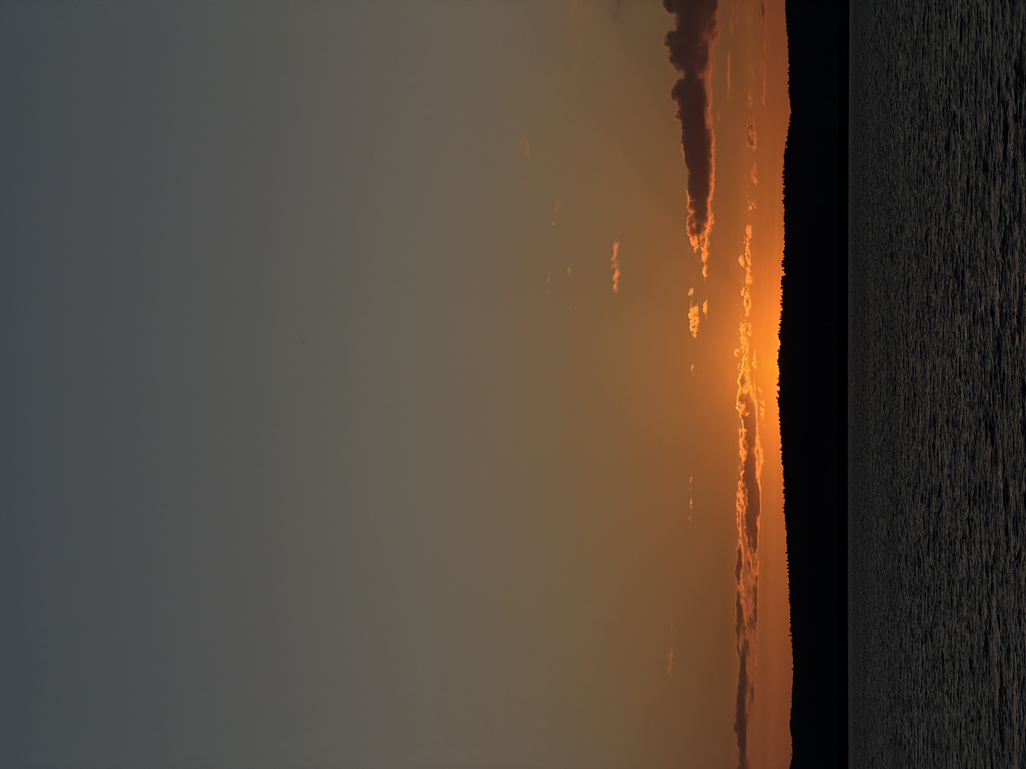 The image depicts a serene sunset over the Kama River in Chaikovsky, Russia. The sky transitions from a deep slate gray at the top to a vibrant orange hue near the horizon, illuminated by the setting sun. Scattered clouds, tinged with orange and dark gray, dot the sky.
A dark silhouette of a forested shoreline stretches across the middle ground, with the dense trees forming an unbroken line against the brightly lit sky. Below the shoreline, the calm waters of the Kama River occupy the lower portion of the image. The water is dark and reflects the muted tones of the sky, with subtle ripples suggesting a gentle breeze.
The scene is devoid of any human presence or activity, emphasizing the natural beauty and tranquility of the landscape at dusk. The overall impression is one of peacefulness and the quiet end of a day.