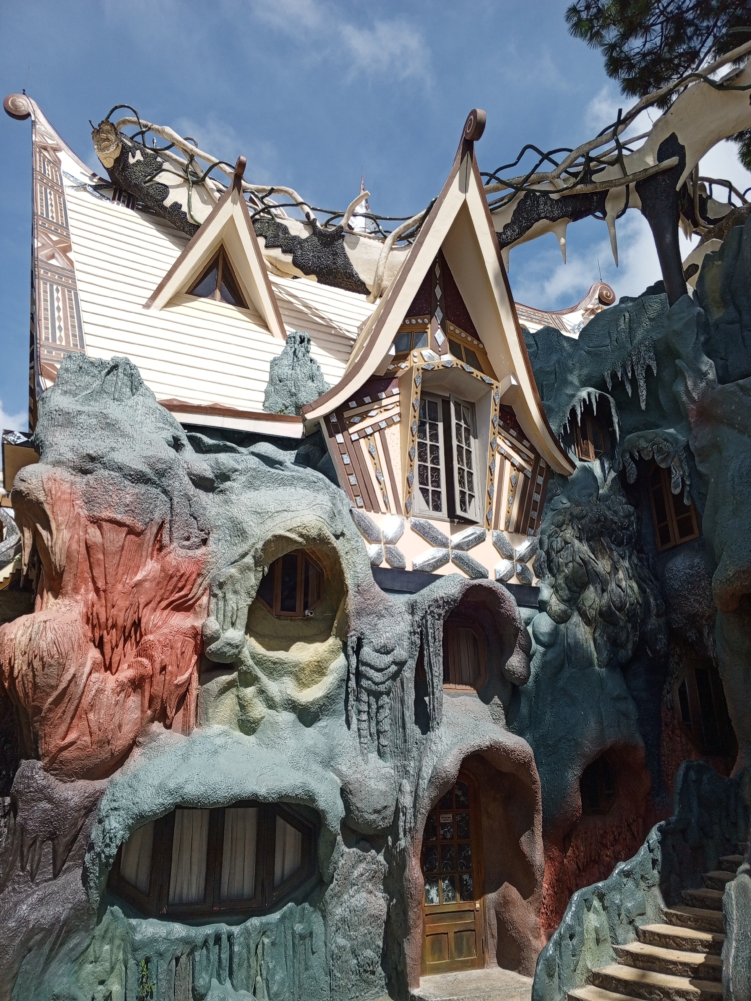 The image depicts the exterior of the "Crazy House" or "Hang Nga Guesthouse" in Da Lat, Vietnam. The building's unconventional architecture resembles organic forms like trees, mushrooms, and caves, with a facade constructed from rough, sculpted concrete in various earthy tones and textures. The main structure, visible at the top, is a multi-story building with a white clapboard exterior and uniquely shaped brown and white patterned rooflines. It features several windows, including a prominent one with an open sash. The surrounding "rock" formations are elaborately molded and painted, creating an illusion of natural caves and grottoes. These formations incorporate arched doorways and small windows, some with wooden frames. A set of stone steps ascends along the right side of the structure. The sky above is blue with scattered white clouds, and a portion of a pine tree branch is visible in the upper right corner. The scene is captured from a slightly low angle, emphasizing the unusual and surreal nature of the architecture. There are no people or animals visible in the image.