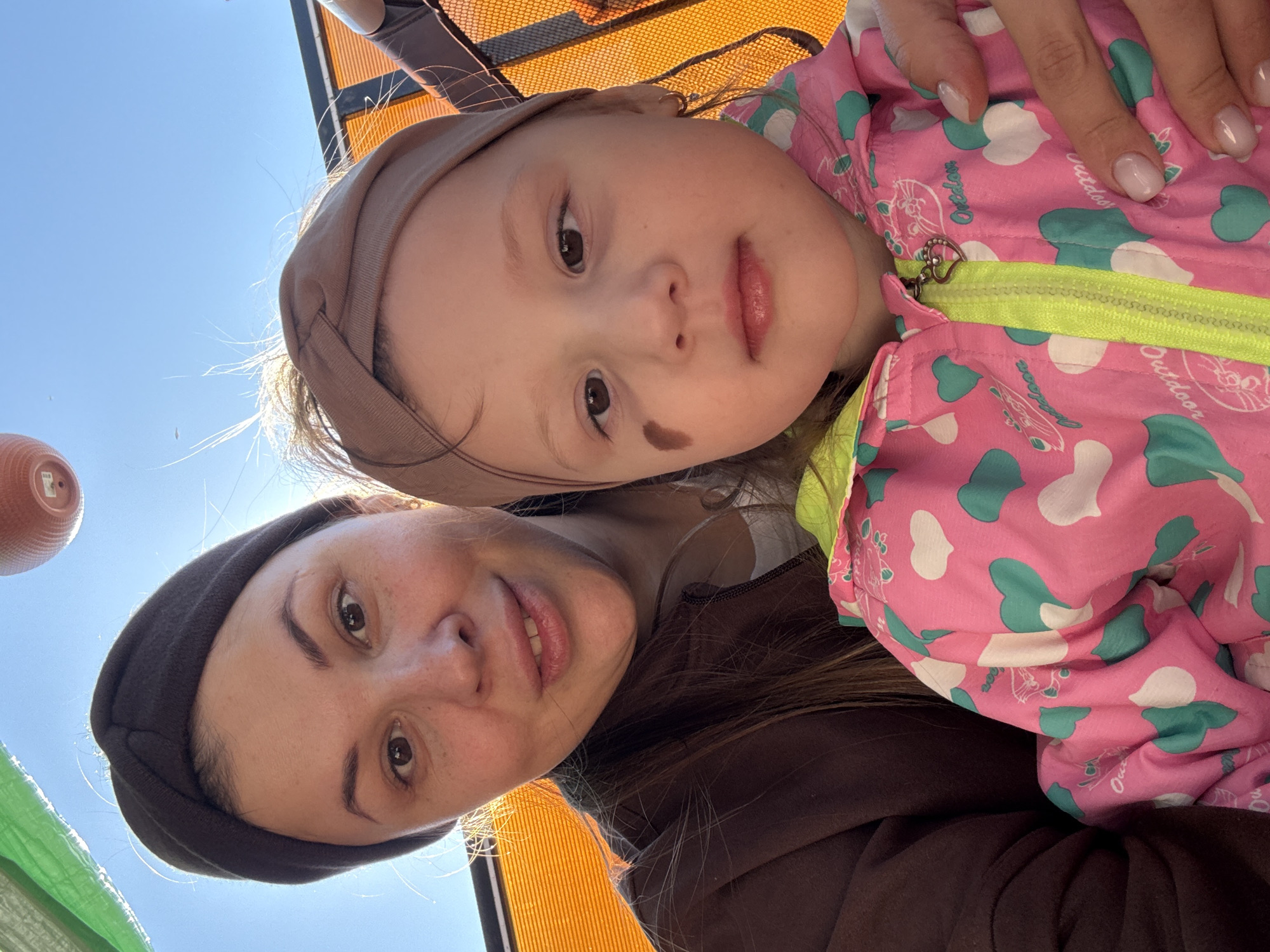 A selfie shows a woman and a toddler, likely outdoors in Кипчак, Russia. The woman wears a dark headband and a brown top. The toddler, resting against the woman, wears a pink jacket with green heart patterns and a light-brown headband. A hand with manicured nails is visible near the toddler's jacket. An orange chair and a blue sky are in the background.