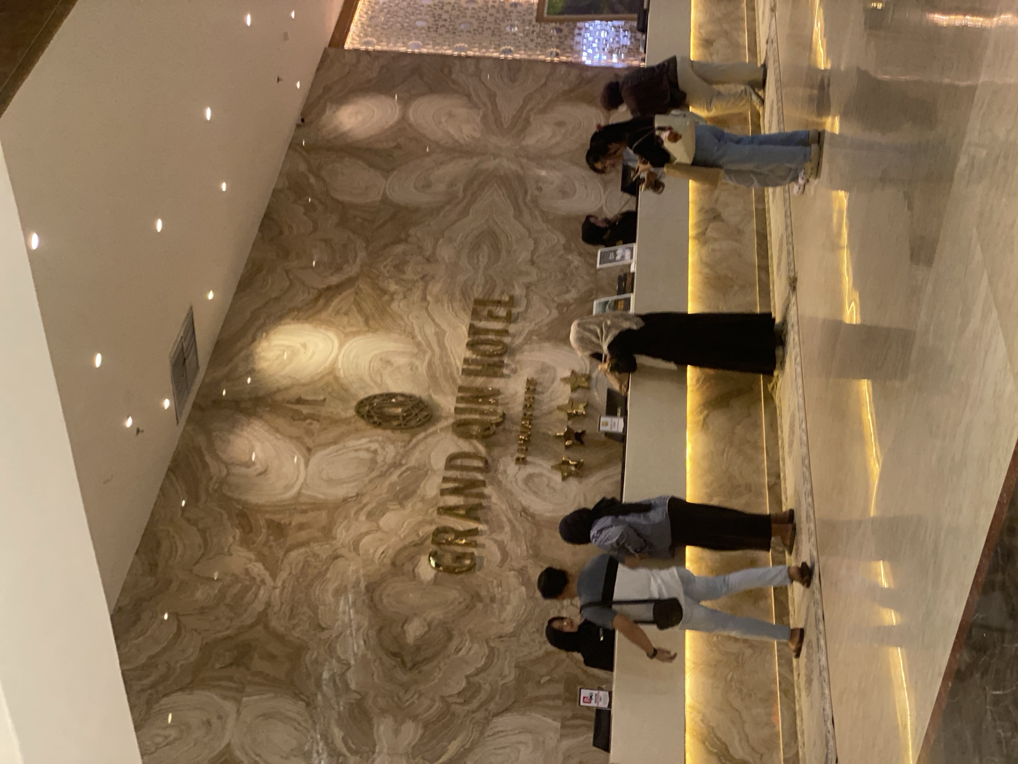 The image shows the lobby of the Grand Q Mall Hotel in Banjarbaru, Indonesia. The hotel's name is displayed in large gold lettering on a marble wall with a swirling pattern. A reception desk is in front of the wall, staffed by several employees in black uniforms. Several guests are interacting with the staff or walking in the lobby. The floor is polished marble, reflecting the warm lighting from the ceiling and accent lights along the reception desk.