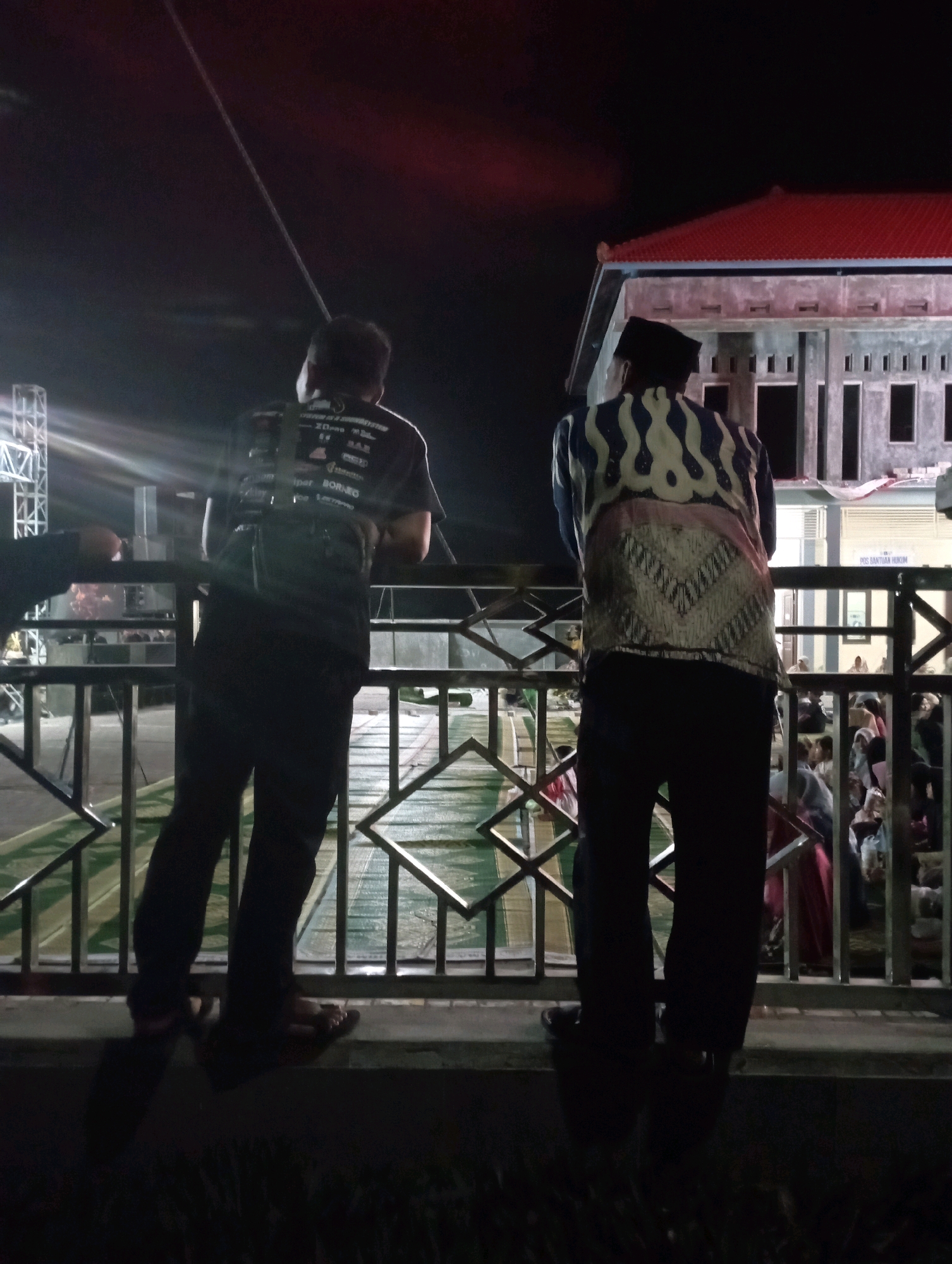 In Ketapang Lor, Indonesia, two individuals are captured in a nighttime outdoor scene. The person on the left, seen from the back, wears a dark t-shirt with text and graphics, dark pants, and sandals. They are positioned behind a metal railing, looking towards a stage with bright lights and equipment. To the right, another individual, also viewed from behind, is wearing a black peci hat, a patterned shirt with blue and white wavy designs on the upper part and a geometric pattern on the lower part, and dark pants. This person is also behind the same metal railing, looking towards a building with a red roof and a light-colored facade. A crowd of people is visible in the background near the building. The setting appears to be an event, possibly a festival or gathering, with stage lighting suggesting a performance or presentation. The overall ambiance is dark, with artificial lights illuminating parts of the scene.