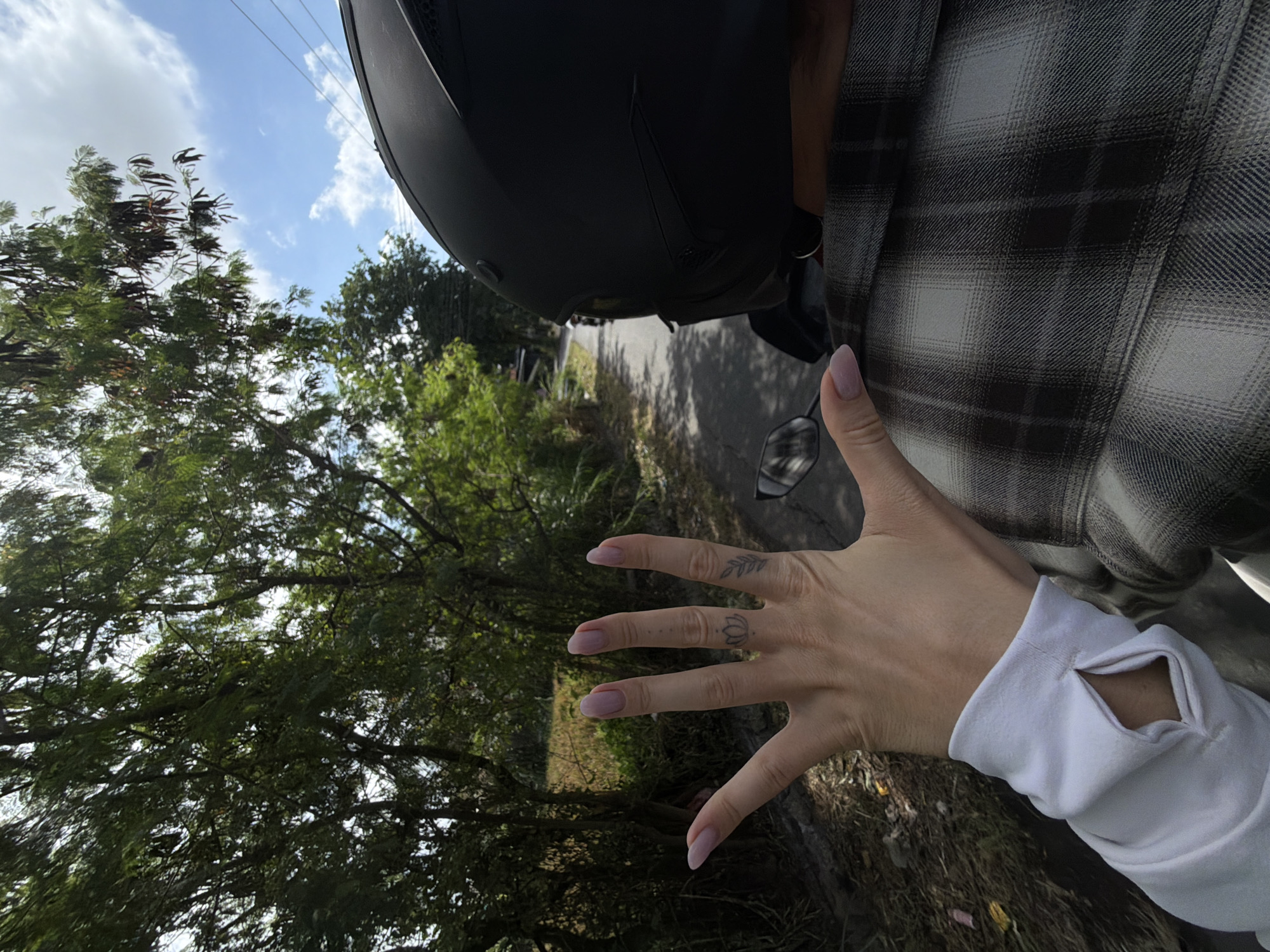 A first-person perspective shot shows a hand with light pink fingernails extended outward, fingers spread, with a lotus tattoo visible on the ring finger and a branch tattoo on the pinky finger. The hand is covered by a white long-sleeved shirt with an opening at the wrist. The person is likely riding on the back of a motorcycle, as indicated by the black helmet visible in the foreground, and a person in a plaid shirt. The background is a rural road lined with trees, with a blue sky and scattered clouds above. The scene appears to be in a tropical or subtropical climate. The location is stated to be in the city of Mengwi, Indonesia.