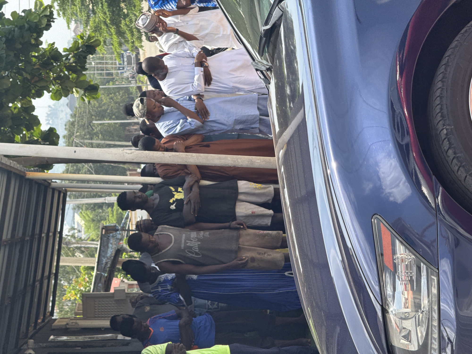 The image shows a group of men lined up, some in traditional clothing and others in casual wear. They appear to be standing in a queue, possibly waiting for something. Some of the men are wearing caps, including a patterned kufi. The background suggests an outdoor setting with trees, foliage, and a fence or wall. A blue car is prominently featured on the right side of the frame, partially obscuring the view of the men. The lighting indicates it is daytime, with sunlight casting shadows. The men's expressions are not clearly discernible, but the overall scene suggests a gathering or event. Some text is visible on a grey t-shirt worn by one of the men, which reads "California". Another man wears a blue and white striped shirt, possibly a soccer jersey. The scene appears to be in a somewhat informal setting, with a corrugated metal structure visible on the left. The presence of traditional attire alongside modern clothing suggests a diverse group of individuals. The image is oriented vertically.