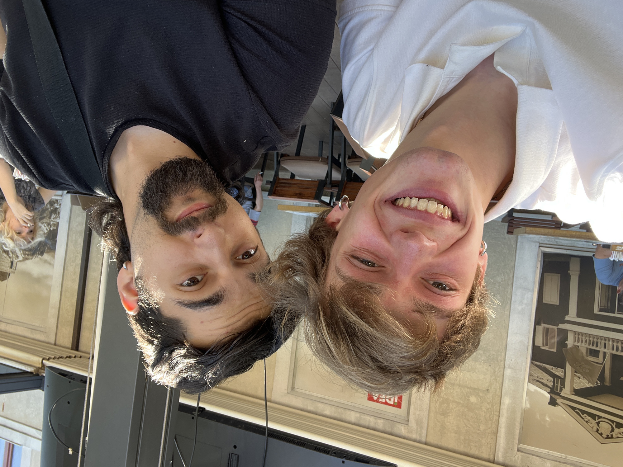 The image is a selfie taken outdoors, likely on a sunny day, featuring two young men. The man on the left has light brown hair and is smiling widely, showing his teeth. He is wearing a white polo shirt. The man on the right has dark brown hair and a beard, and he is looking towards the camera. He is wearing a black t-shirt. Behind them, there are tables and chairs, suggesting an outdoor seating area of a cafe or restaurant. A building with a mural of a house is visible in the background. The overall impression is that of a casual, social gathering. The setting appears to be the city of Cetinje, Montenegro, given the context provided.