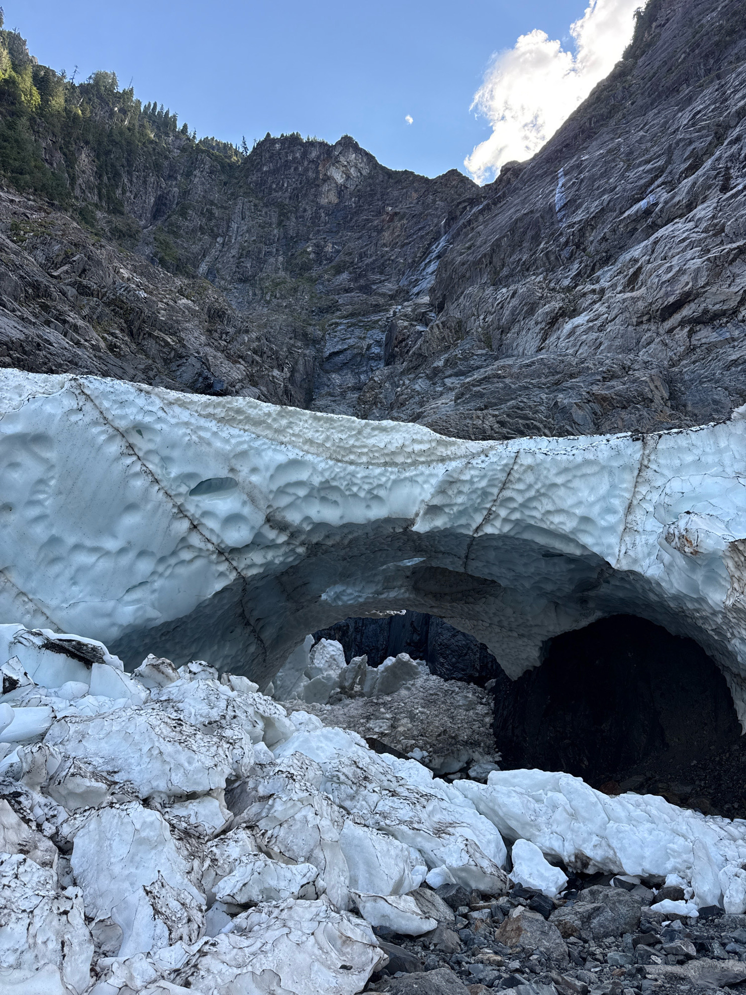 The image depicts a glacial cave entrance set against a backdrop of steep, rocky mountainside with sparse vegetation and patches of snow. The cave opening is framed by a large overhang of textured, blue-tinged ice, with a chaotic pile of icy debris in the foreground. The mountain face above the ice exhibits distinct layers and fissures, with what appears to be a thin waterfall or streaking water running down its surface. The sky is clear blue with a few wispy white clouds, suggesting daylight. The location is identified as Silverton, United States, though specific landmarks within the city are not discernible from the image. There are no people or animals visible in the scene.