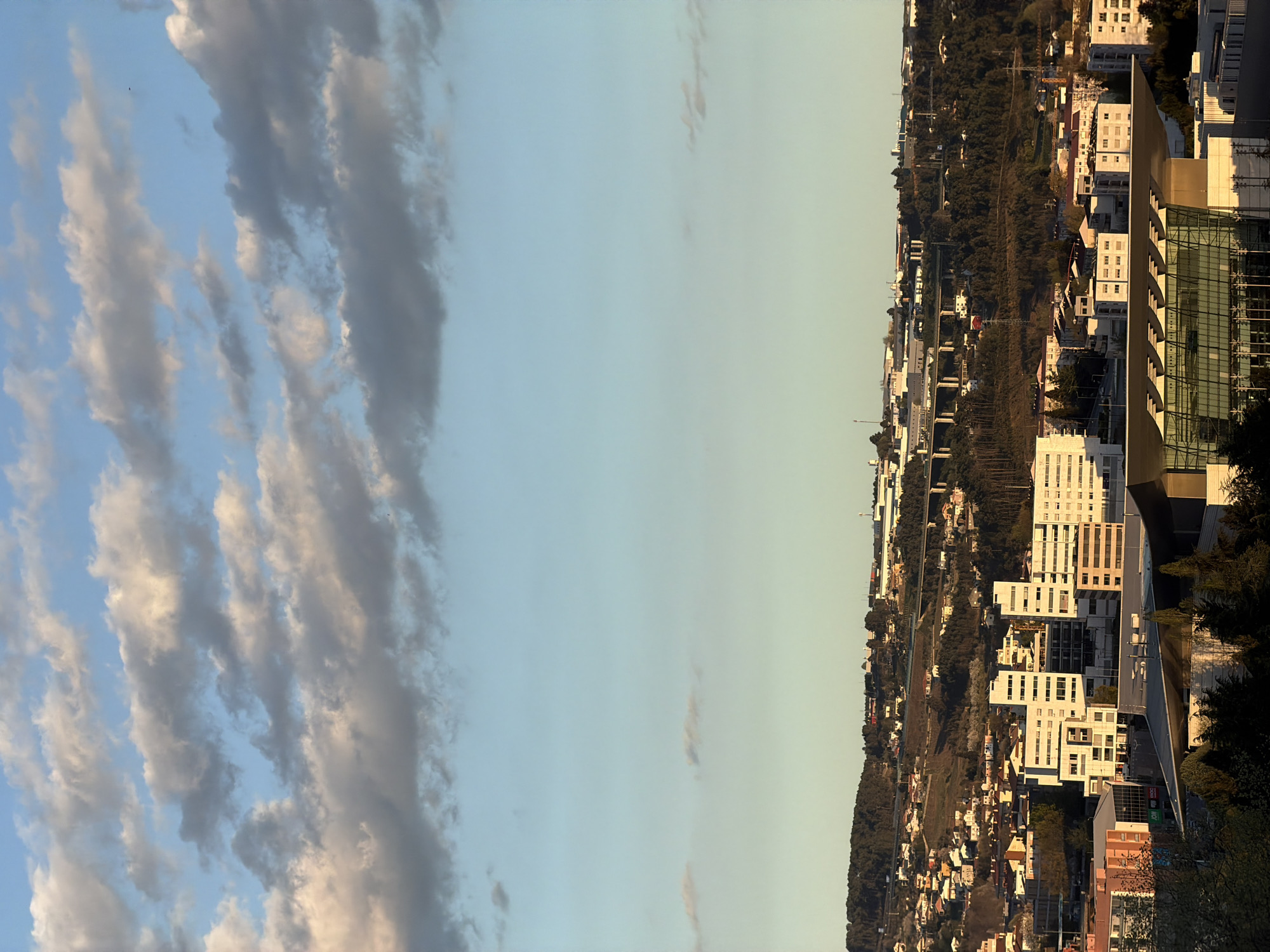 The image shows a cityscape in Vila Real, Portugal, viewed from an elevated position. The city is situated on a hilly terrain, with a mix of residential and commercial buildings. Modern white apartment complexes contrast with traditional houses and dense vegetation. A prominent building with a glass facade stands out amidst the others. The sky is predominantly clear with patches of altostratus clouds with subtle shading. There is no visible human activity.