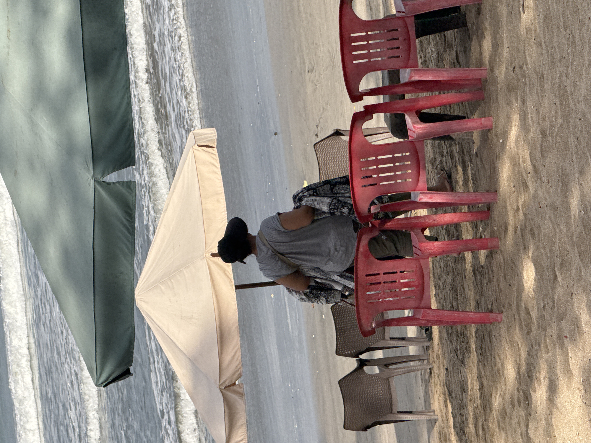 A person wearing a hat and a grey t-shirt is standing on a sandy beach in Kuta, Indonesia. They are holding a patterned cloth. Two beach umbrellas are set up, one light-colored and one dark green. In the foreground, there are several plastic chairs, some red and some brown. The ocean waves are visible in the background.