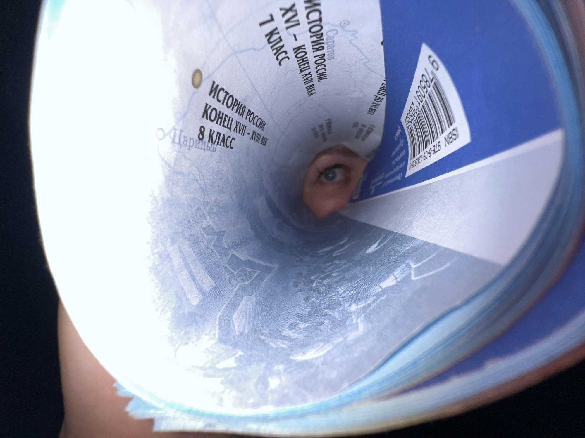 A close-up, angled view from within a tightly rolled collection of papers or books reveals a human eye looking directly towards the viewer. The eye, positioned centrally within the cylindrical opening, has a light iris. The rolled material consists of multiple pages, primarily light blue and white, featuring Cyrillic text and map-like graphics. Visible text fragments include "ИСТОРИЯ РОССИИ. XVI – КОНЕЦ XVII ВЕКА. 7 КЛАСС" (History of Russia. XVI – late XVII century. 7th Grade), "ИСТОРИЯ РОССИИ. КОНЕЦ XVII – XVIII ВЕК. 8 КЛАСС" (History of Russia. Late XVII – XVIII century. 8th Grade), and city names such as "Саратов" (Saratov) and "Царицын" (Tsaritsyn). An outer, darker blue cover of one of the books displays a barcode and the ISBN "978-5-09-120535-0". The edges of the rolled material are brightly illuminated, creating a stark contrast with the dark background surrounding the object. A faint skin tone is visible on the left, suggesting a hand is holding the rolled item. The specific location in the city of Noviy, Russia, is not discernible from the image; the visible textual content pertains to general Russian history and specific historical cities within Russia.