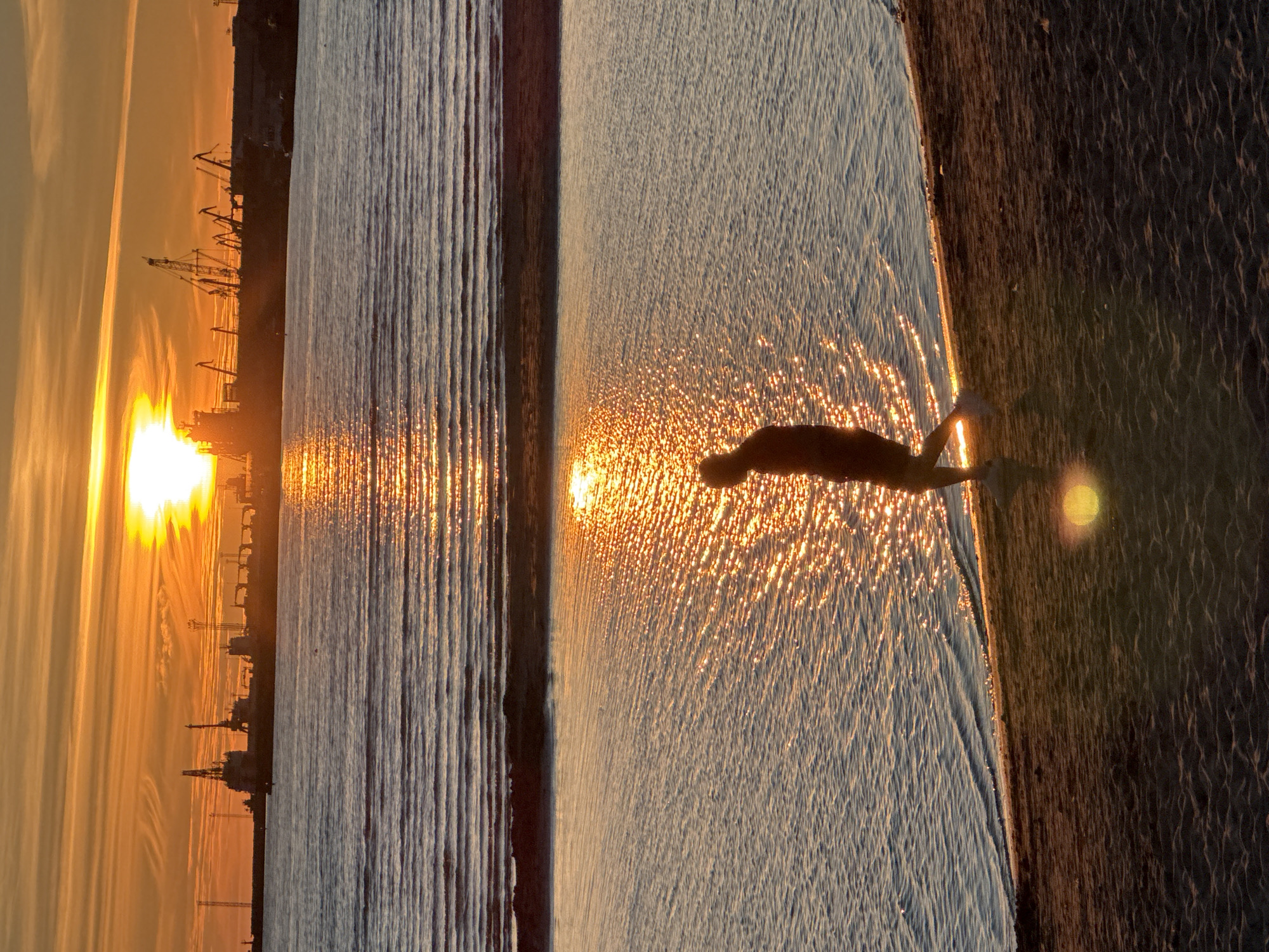 The image shows a sunset over a harbor in Tallinn, Estonia. The sun is a bright orange ball low on the horizon, casting a golden glow across the sky and the water. The water is calm with gentle ripples, reflecting the warm colors of the sunset. In the foreground, a person is walking along a sandy beach towards the right of the frame. They are silhouetted against the bright water, and appear to be wearing shorts and sneakers. The beach itself is dark and sandy, with some texture visible. In the background, the silhouette of the Tallinn harbor is visible, with several large ships and cranes standing out against the colorful sky. The sky is filled with streaks of orange, yellow, and pink clouds.