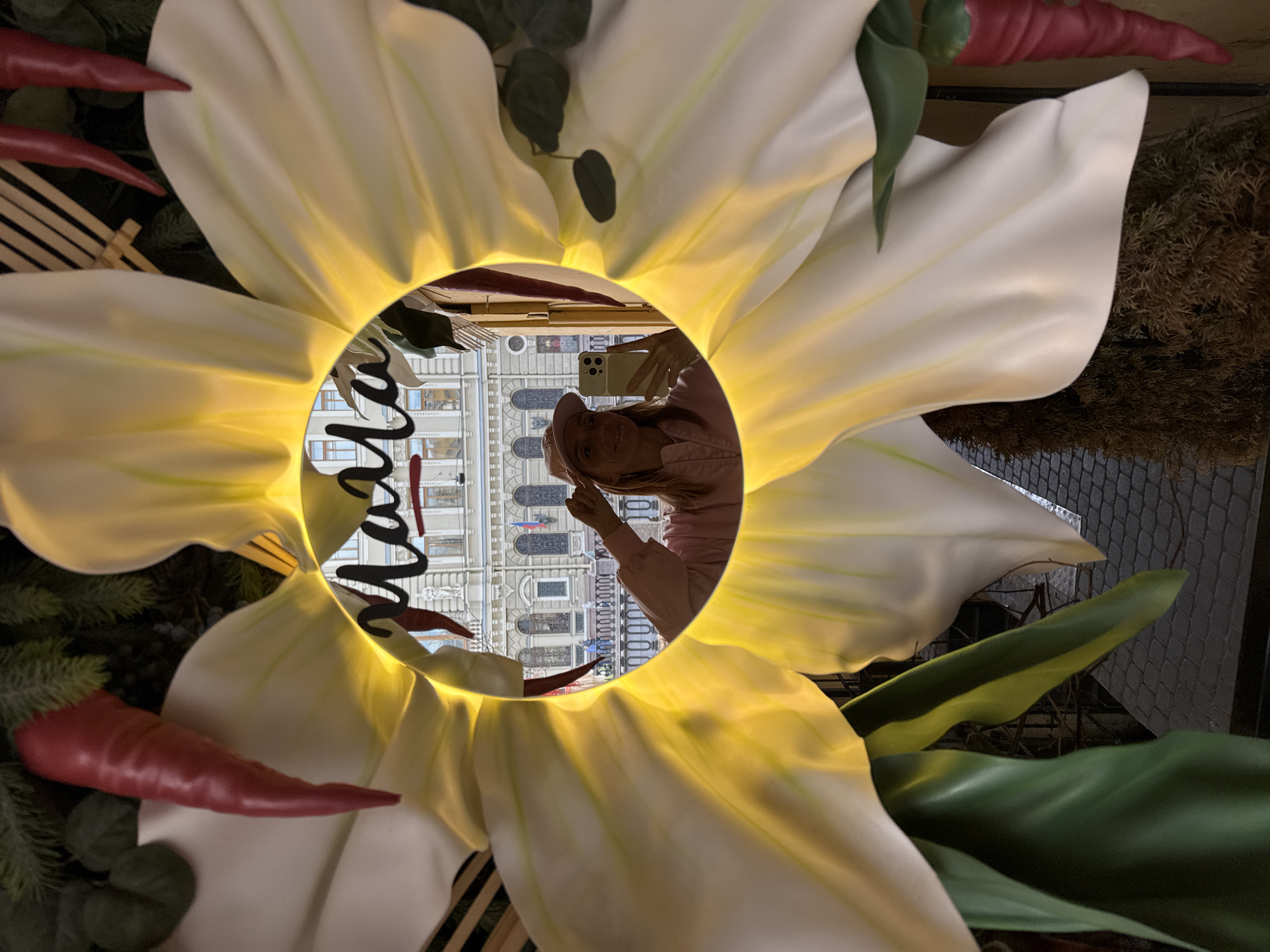 The image shows a reflection of a woman in a flower-shaped mirror, likely a decorative installation in Saint Petersburg, Russia. The woman is holding a smartphone, possibly taking a photo or video. The reflection also captures a portion of the building in the background, revealing architecture characteristic of Saint Petersburg, with multiple windows and ornamentation. Additional decorative elements, including carrots, are visible around the flower mirror.