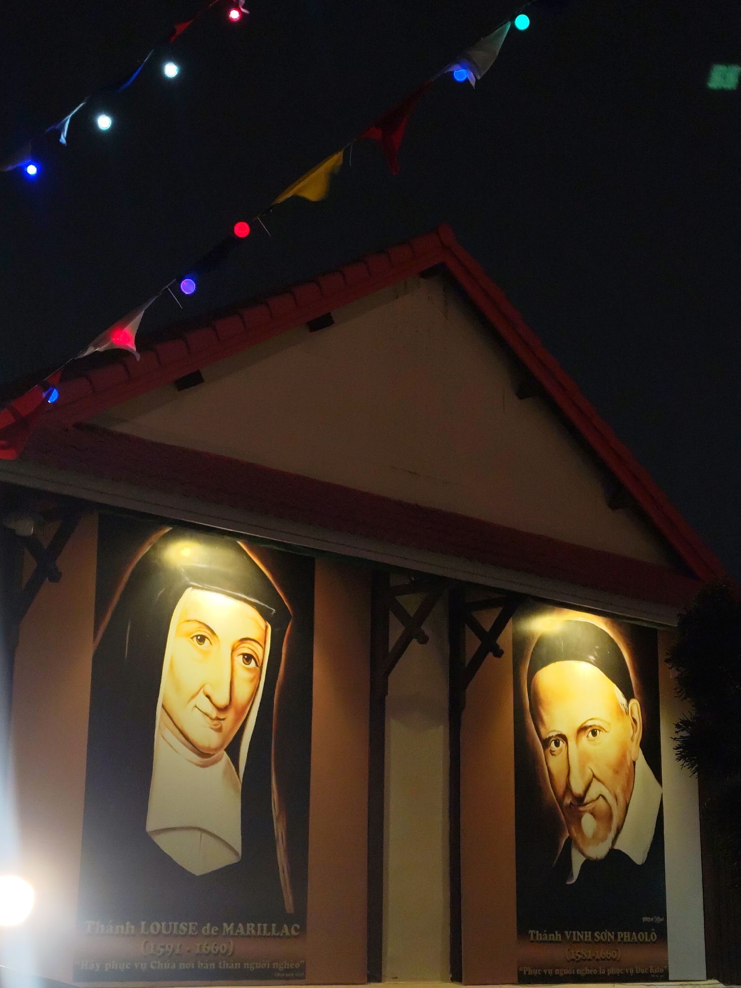 The image captures the exterior of a building in what is identified through text on the walls as likely being in Da Lat, Vietnam. The building's facade features paintings of what appear to be religious figures: Saint Louise de Marillac and Saint Vincent de Paul, identifiable through inscriptions in Vietnamese. Above the building, a string of multi-colored lights drapes across the sky, creating a festive or celebratory atmosphere. The scene seems to be set at night, with a dark sky providing a contrasting backdrop to the illuminated paintings and decorations. The building's architectural design includes a pitched roof with visible eaves. Part of a tree is also visible to the right of the building.