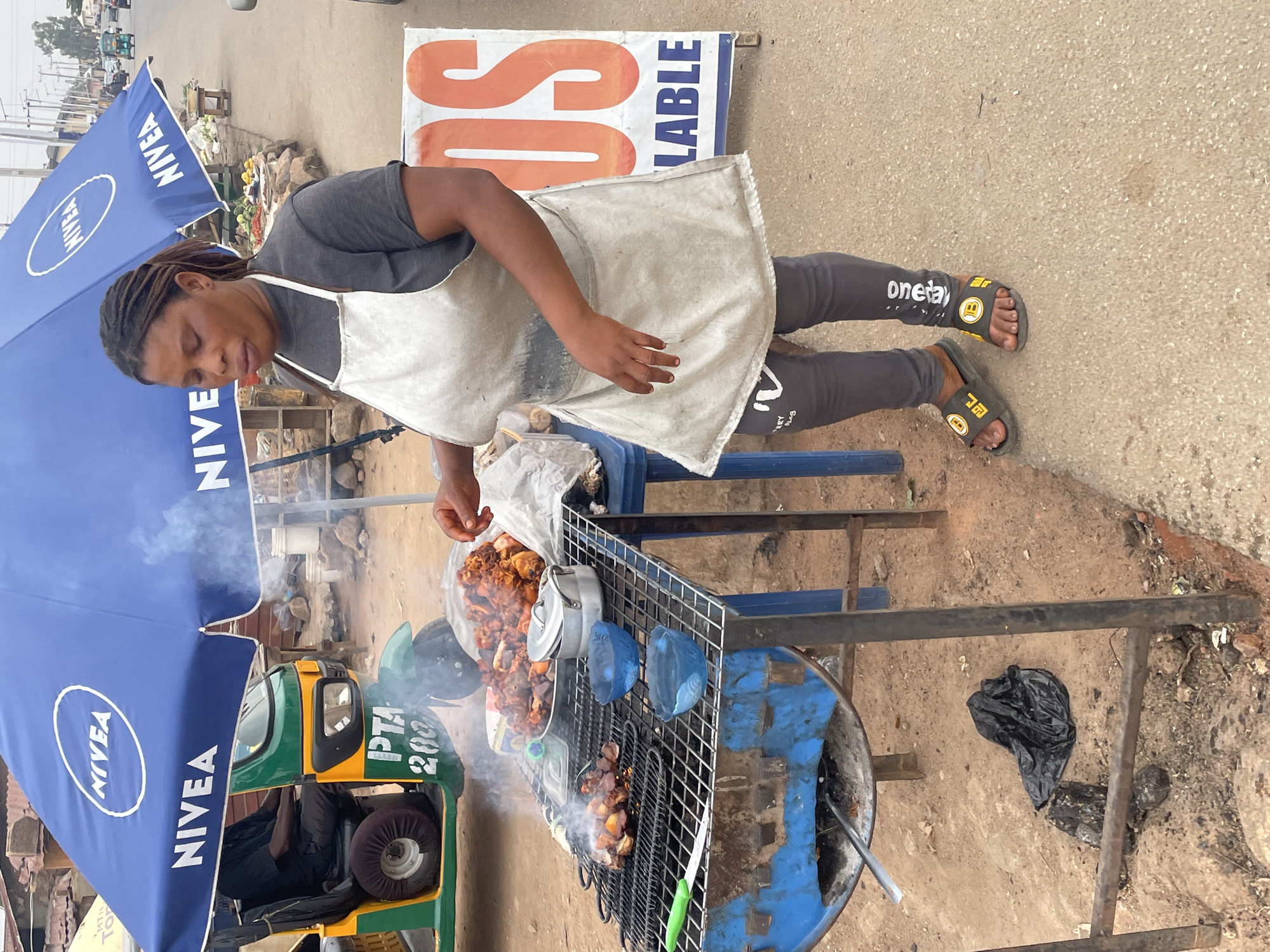 In Jos, Nigeria, a woman wearing a gray t-shirt, gray leggings with "oneday" printed on the leg, and a white apron is standing next to a metal grill, cooking food over a blue barrel. Smoke rises from the grill. She wears sandals. A large blue umbrella with "NIVEA" printed on it provides shade. A green and yellow three-wheeled vehicle is partially visible under the umbrella. A sign with "SOS AVAILABLE" is in the background. A black plastic bag is on the ground nearby. A plate of cooked food sits to the side of the grill.