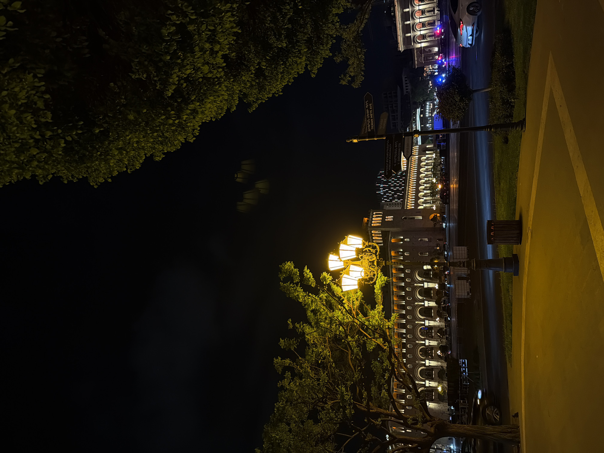 This nighttime image depicts a bustling street scene in Yerevan, Armenia, illuminated by artificial lights. The perspective is slightly elevated, looking down a street lined with buildings and trees.
In the foreground and to the left, dark green foliage from trees partially fills the frame, with some leaves catching the golden light from streetlamps. A prominent, ornate streetlamp with multiple glowing yellow lanterns stands in the midground, its light casting a warm glow on the surrounding area and a large tree next to it.
The street, which appears damp and reflective, runs parallel to a series of multi-story buildings. These buildings showcase distinctive architecture with multiple arched windows and balconies, many of which are illuminated, suggesting commercial establishments or residential spaces. A white car with flashing blue and red emergency lights is visible on the street, parked or slowly moving. Another vehicle is partially visible further down. A dark trash bin is positioned on the right side of the street near a light-colored paved area, possibly a sidewalk or another building facade.
A street signpost with multiple directional arrows is visible, providing location cues. The visible text from the signs includes "ԵՐԵՎԱՆԻ ՔԱՂԱՔԱՊԵՏԱՐԱՆ" (YEREVAN MUNICIPALITY), "Վ. ՍԱՐԳՍՅԱՆ ՓՈՂ" (V. SARGSYAN STR.), and "ՏԻԳՐԱՆ ՄԵԾԻ ՊՈՂ" (TIGRAN METS AVE.). Another sign reads "VERMISSACE," likely a business name. The sky above is dark, confirming the late hour. The scene captures the lively ambiance of a city at night.