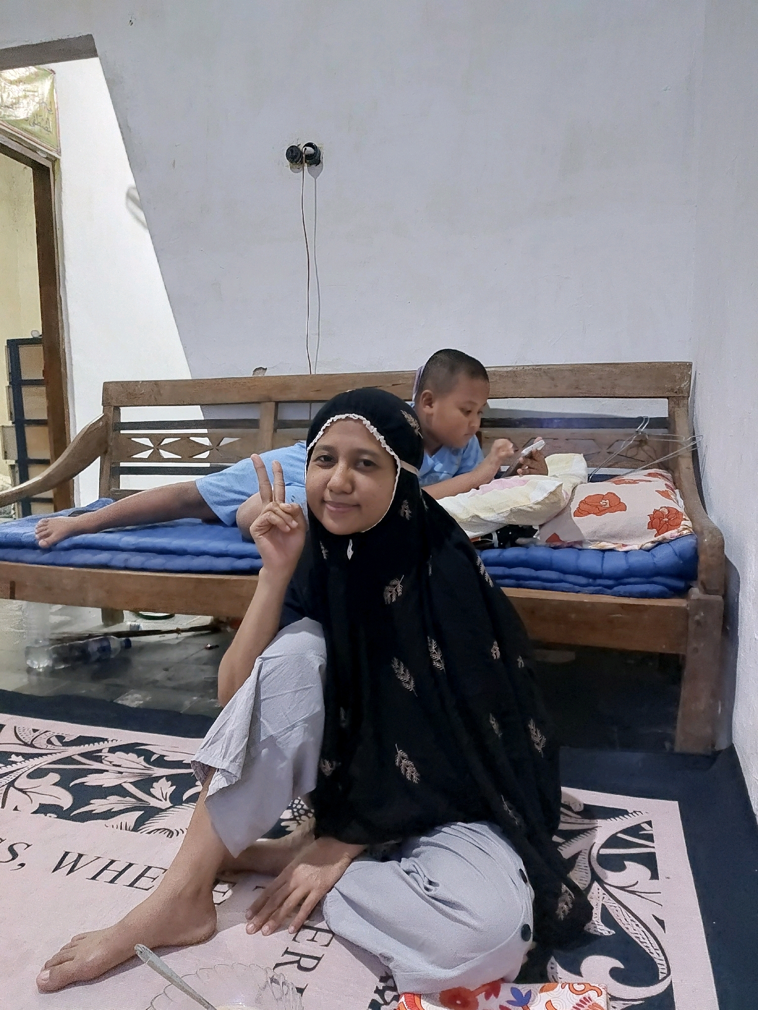 In Sarangan, Indonesia, a woman wearing a black hijab and grey pants is sitting on a rug with a patterned design in the foreground. She is making a peace sign with her right hand. Behind her, a young boy in a blue t-shirt is lying on a wooden bench with a blue mattress and pillows. He appears to be looking at a mobile phone. The scene is indoors, with a white wall and a wooden structure visible in the background. A plastic bottle of water and a spoon are on the floor near the woman.
