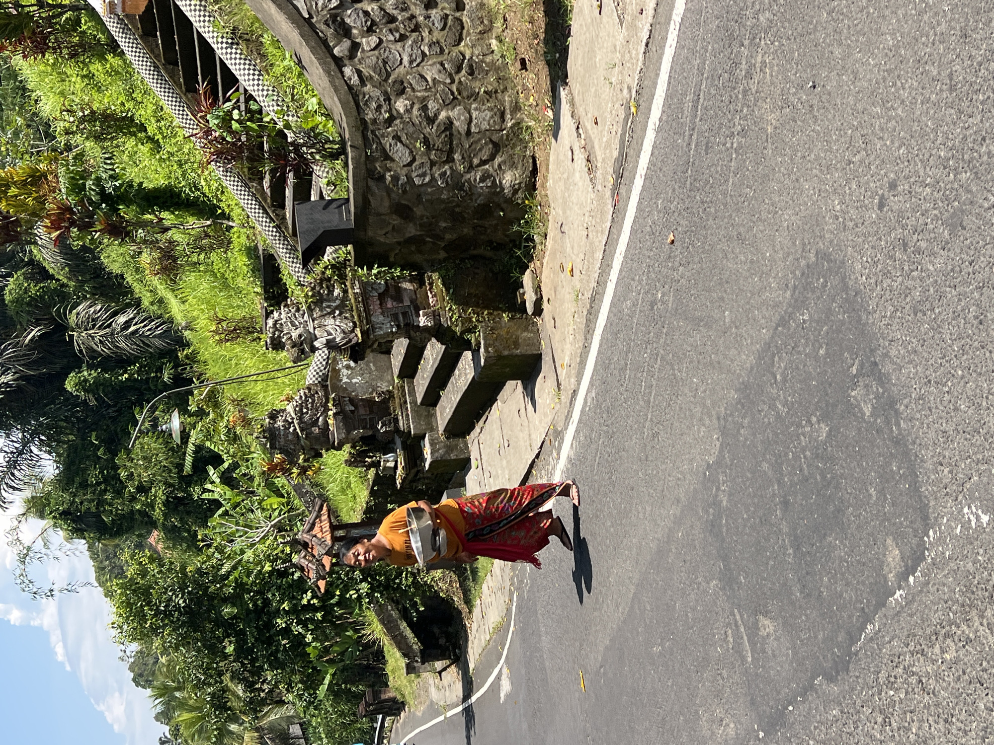 A mid-shot depicts a woman walking on the edge of an asphalt road in Sembung, Indonesia. The woman, appearing middle-aged, wears an orange short-sleeved shirt, a red and brown patterned sarong, and brown sandals. She holds a silver metal basin with both hands at waist level and smiles towards the viewer. A distinct shadow is cast to her left on the road surface. The dark gray asphalt road features a faded white solid line marking its edge. To the right, a light gray concrete sidewalk leads to a greyish-brown stone retaining wall. Stone steps ascend the embankment, flanked by balustrades decorated with black and white checkered patterns. Ornate stone structures, possibly shrines, are visible at the base of the stairs. The embankment and background are densely covered with lush tropical vegetation, including palm trees, banana plants, and various shrubs. A curved street lamp with a green shade is visible amidst the foliage on the upper right. The sky above is partially visible, showing clear blue with some white clouds.