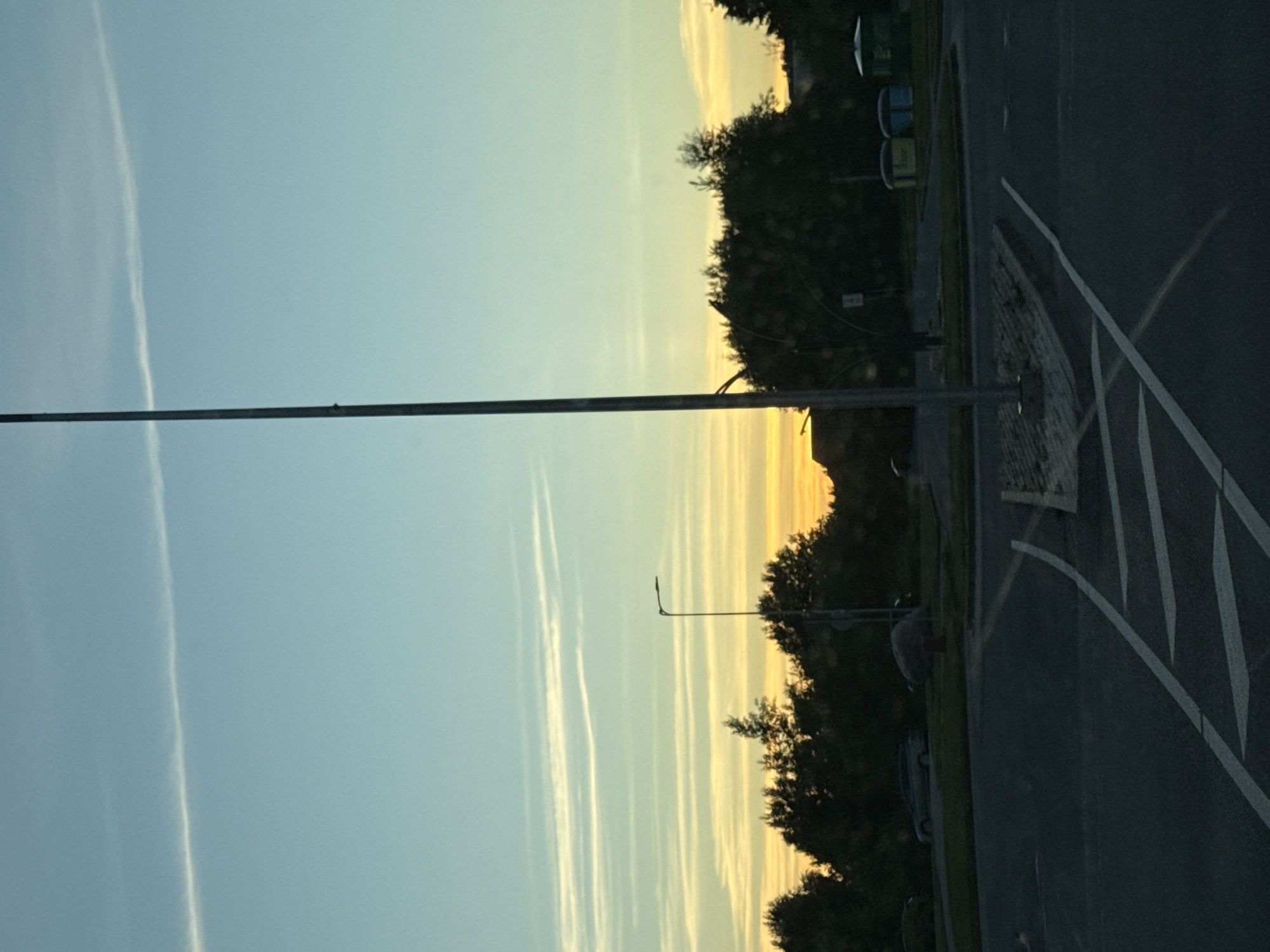 A wide, low-angle photograph shows a street scene at dusk, possibly in Tallinn, Estonia. The sky is a gradient of pale blue at the top, transitioning to yellow and orange near the horizon. Wispy clouds streak across the sky. In the foreground, a paved road with white markings is visible, suggesting a junction or parking area. A tall, slender lamppost stands prominently in the center of the frame. To the left of the lamppost, trees form a dark line against the lighter sky. In the mid-ground, several cars are parked on the grass verge. Further back, two brightly colored portable toilets are visible. The overall impression is one of a quiet, suburban or commercial area at the end of the day.