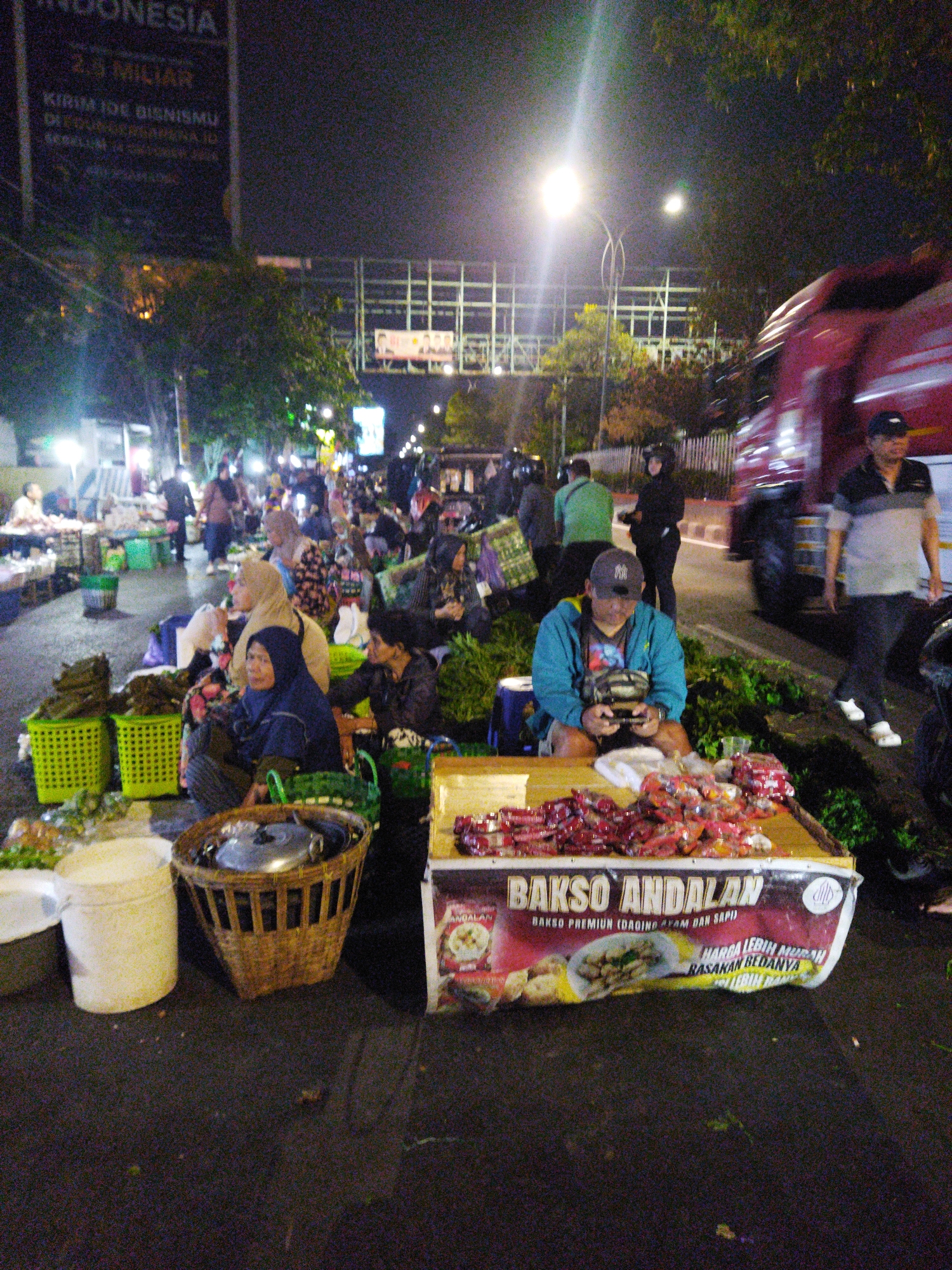 This is a nighttime street scene in Semarang, Indonesia, likely a bustling market area. In the foreground, a man wearing a blue jacket and a black baseball cap sits on a wooden cart filled with pre-packaged red food items. He is looking down at his hands, holding what appears to be a mobile phone or wallet. Beside him, a woven basket rests on the ground, containing a metal pot lid. Behind the cart, two women are visible, one wearing a blue headscarf and the other a beige headscarf, both appearing to be engaged in conversation or tending to their goods.
Further back, numerous people are scattered throughout the scene, some walking, others gathered around stalls. Various market goods, including vegetables and plastic baskets, are visible. A large red truck is partially in frame on the right side of the image, its lights illuminating the scene. In the background, streetlights and overhead structures suggest an urban environment. The overall impression is a lively night market atmosphere.