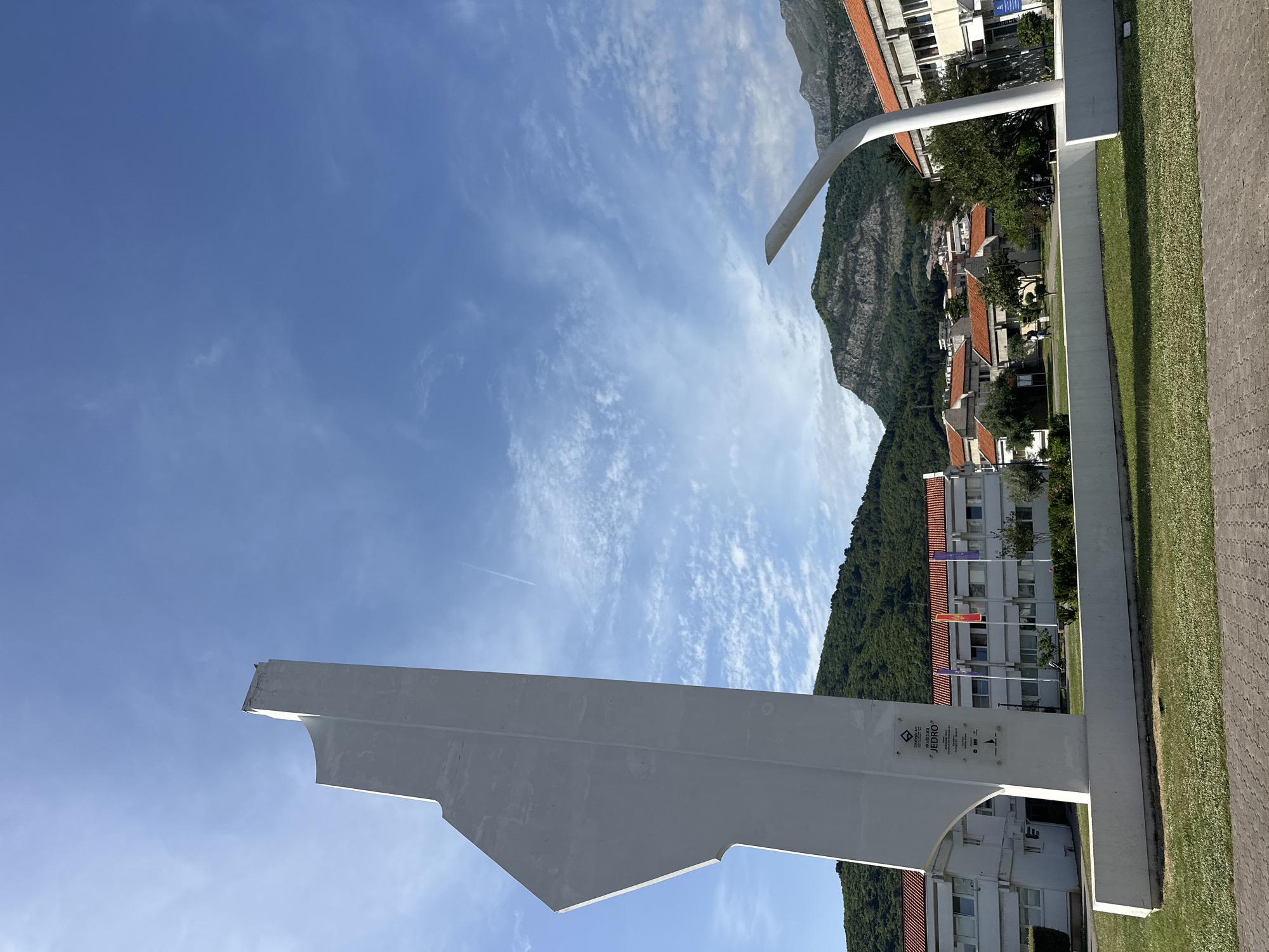 The image shows a daytime view of Budva, Montenegro. In the foreground is a white, angular monument, possibly a public art piece, with visible text "JEDRO" on it. Behind the monument are buildings with red tile roofs and a tree-covered mountain. The sky is blue with scattered cirrus clouds. A contrail from a passing aircraft is visible in the sky. The monument sits on a grassy area adjacent to a paved walkway. No people are immediately visible.