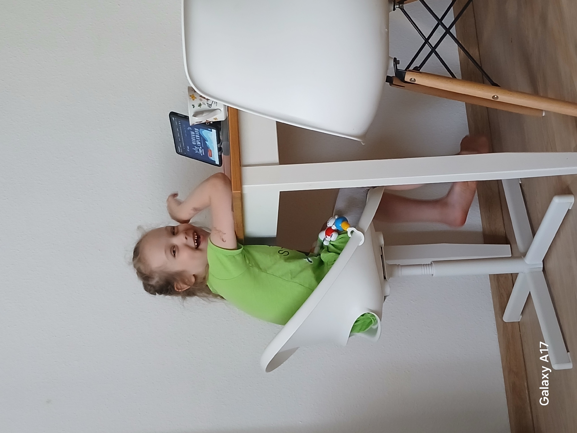A young child is sitting in a white high chair at a table. The child is wearing a green shirt. A smartphone is propped up on the table, displaying a video. The table is next to a white and wood chair. The photo was taken in an indoor location. The city of Yekaterinburg, Russia, is not directly observable.