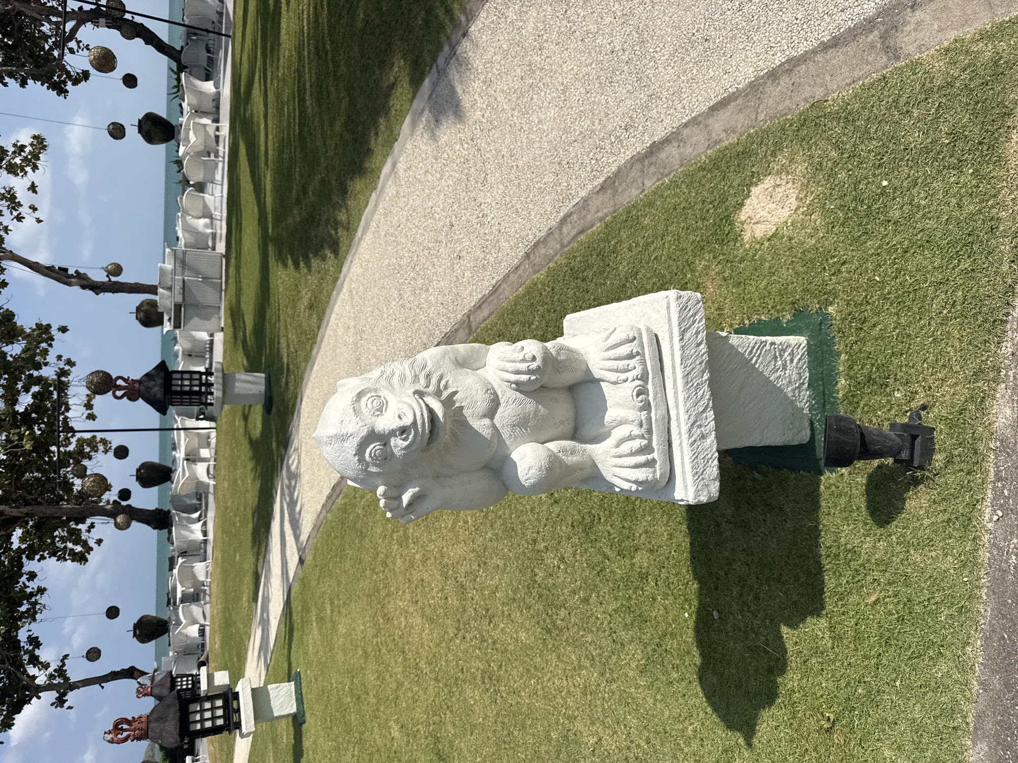 A weathered, light gray stone statue of a monkey sits on a two-tiered pedestal in a grassy area. The monkey is in a seated position, facing forward with its hands raised, and appears to be waving. The statue is positioned on a section of green turf bordered by a light-colored, pebble-textured pathway. In the background, trees with hanging spherical decorations cast shadows on the lawn, and beyond them, the turquoise ocean meets the horizon under a pale blue sky with scattered clouds. A row of white chairs and tables is visible in front of the water, suggesting an outdoor dining or seating area. Several dark, lantern-like structures with ornate tops are also present along the pathway, near the edge of the lawn. The scene appears to be a resort or public space in Kuta Selatan, Indonesia, given the tropical vegetation and coastal setting.
