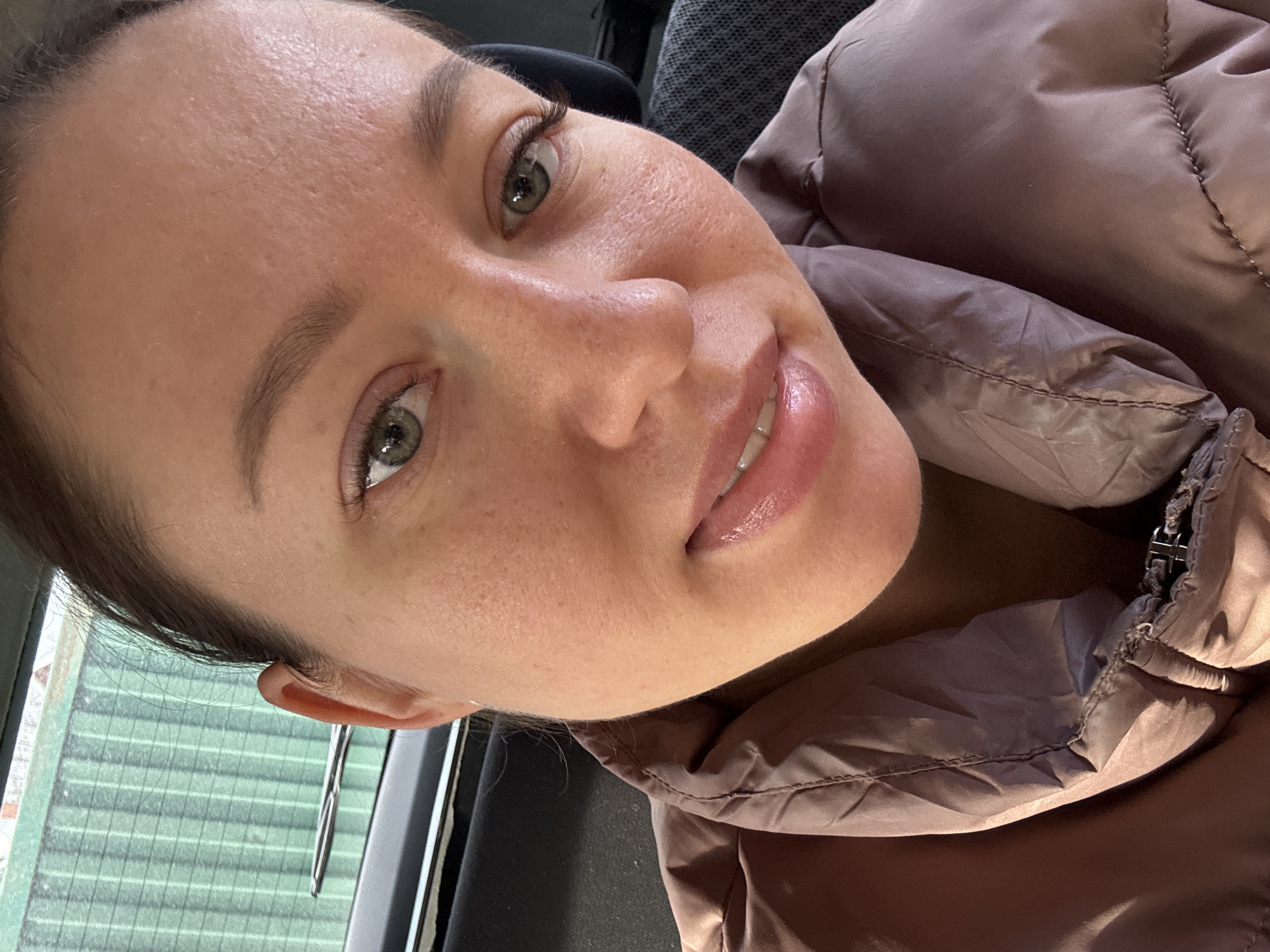 A close-up, front-facing image depicts an adult female subject positioned inside a vehicle. The subject has light brown hair pulled back, green eyes, and is looking directly at the camera with a slight smile, revealing glossy lips. She is wearing a light brown, possibly mauve, puffer jacket with visible stitching. Behind the subject's head, the top of a dark car seat headrest is visible on the right. Through the vehicle's rear window, a green, vertically ribbed metal fence or structure is discernible, along with a portion of a rear windshield wiper blade. The scene is located in the city of Ферма, Russia.