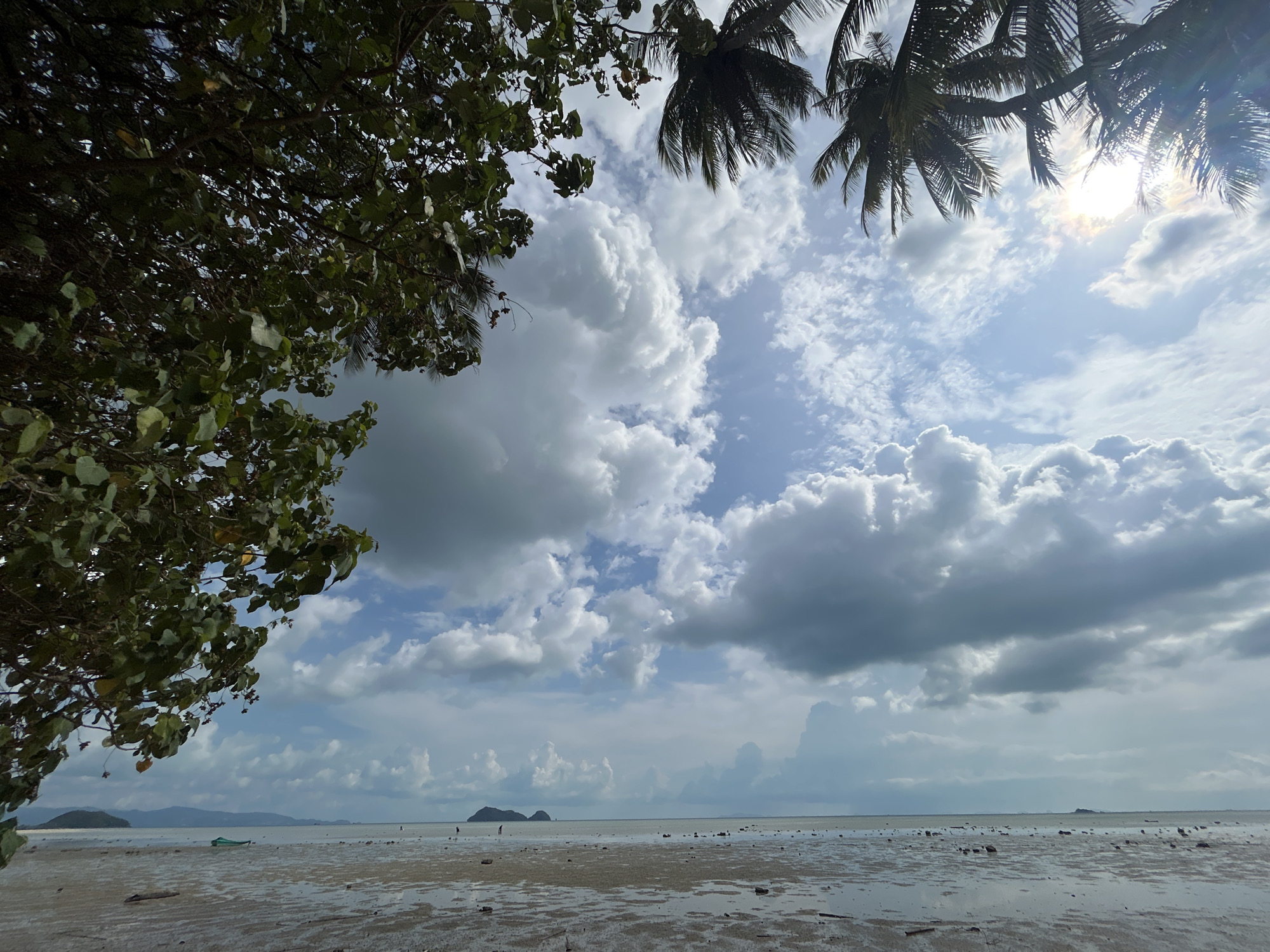 The image depicts a coastal scene at low tide, possibly in or near Baan Sri Thanu, Thailand, given the tropical vegetation and coastal features. The foreground is a muddy tidal flat with scattered rocks and pools of water reflecting the sky. In the mid-ground, the water recedes, revealing more of the tidal flat. Two figures are walking in the distance. A small boat is beached on the left.
The sky is predominantly filled with white and gray cumulus clouds, with patches of blue sky visible. The sun is partially obscured by the palm trees in the upper right corner, creating a lens flare effect. Tree branches with green leaves frame the left side of the image. Other islands are present at the horizon.