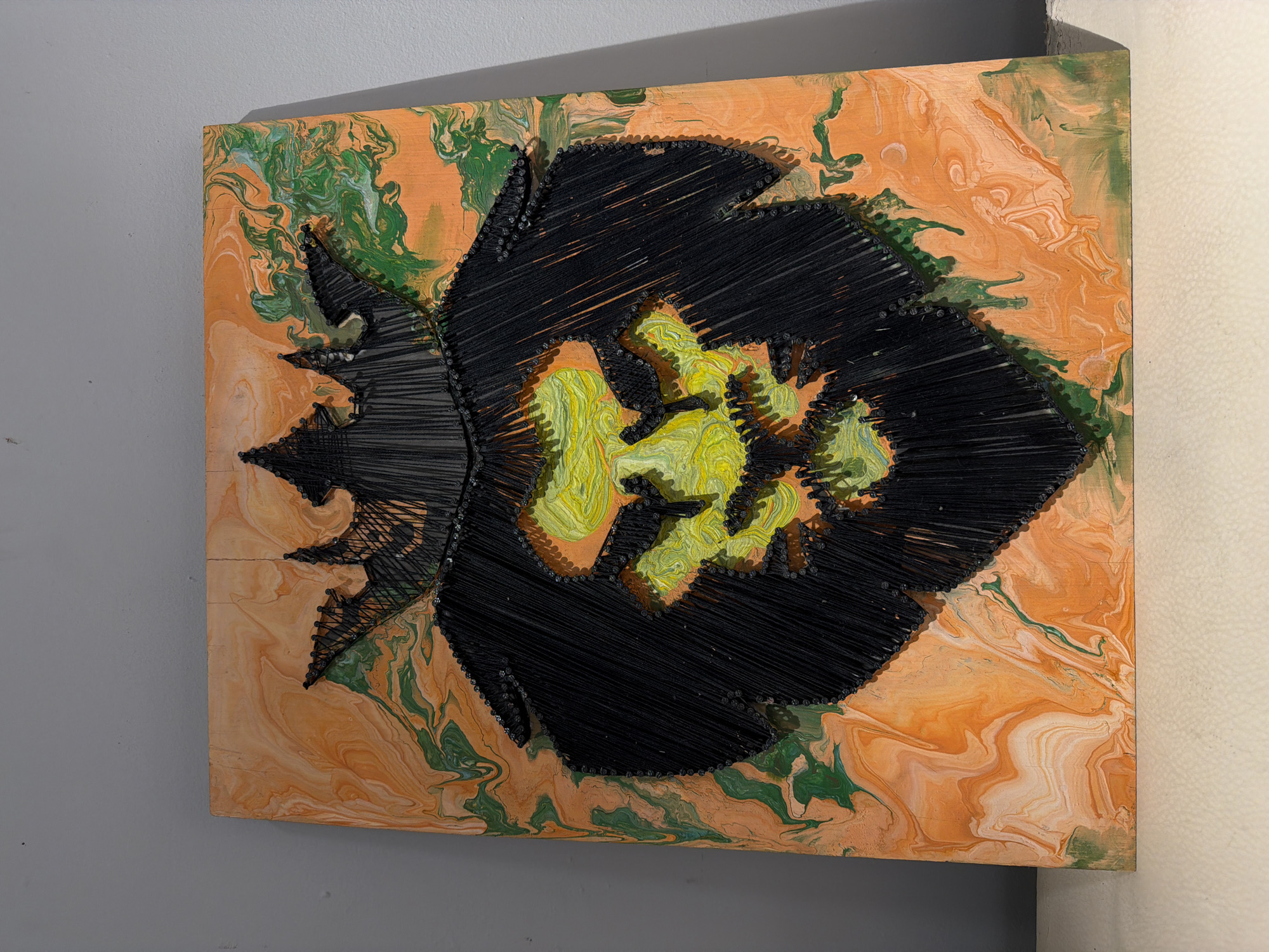 The image shows a piece of abstract artwork. The artwork is a square-shaped piece, mounted on a wall with a light gray surface. The background of the artwork itself features a marbled effect in shades of orange and green. A design resembling a stylized flower is created in the center, outlined with black string connected to nails. The core of the flower is filled with textured yellow and green paint. The artwork is situated indoors. There's no information available about the image's location in Beryozovka, Russia.