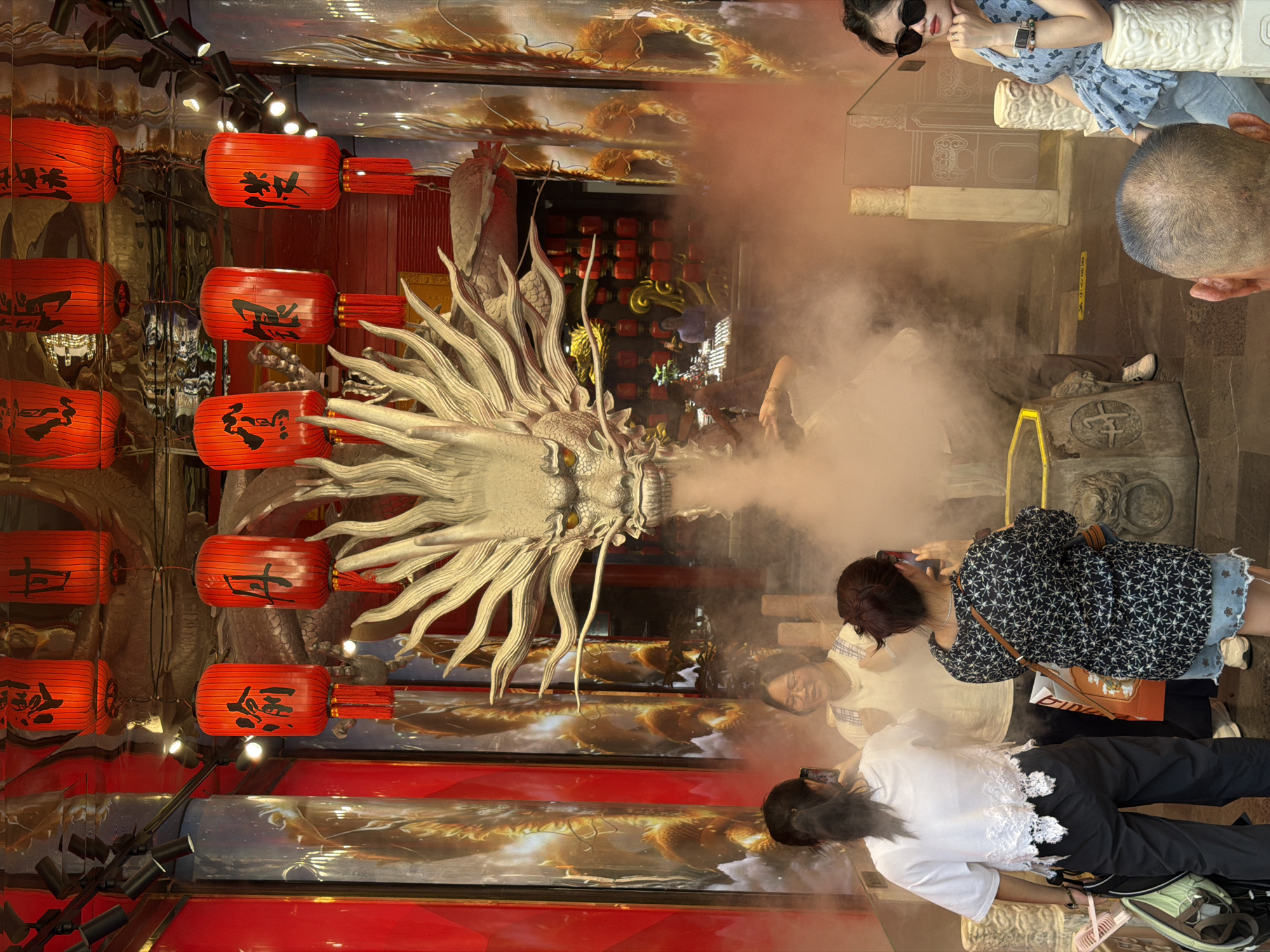 In the Yuzhong District of China, this image captures an indoor scene with a prominent, large, white dragon sculpture spewing smoke from its mouth. Red lanterns with Chinese characters hang from the ceiling, illuminating the space. Several individuals are present: a woman in a white top and black pants with a backpack, another woman in a white shirt and checkered overalls, and a third woman in a black patterned top and denim shorts. The woman in the black top is taking a photo with her phone. Another person, partially visible, is in the foreground with their back to the camera. The background features tall, cylindrical structures adorned with images of golden dragons. The overall ambiance suggests a cultural or entertainment venue, possibly a themed attraction, with a focus on traditional Chinese motifs.