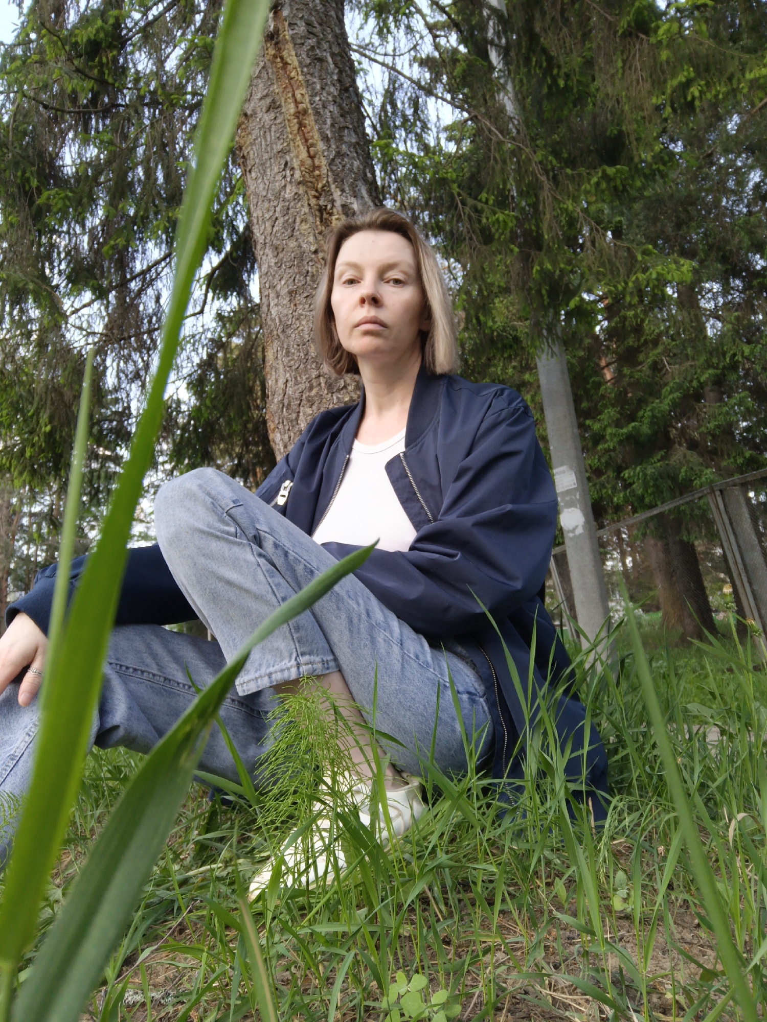 A woman with short blonde hair is sitting cross-legged on the grass in a wooded area. She is wearing a white t-shirt, blue jeans, and a navy blue bomber jacket. She is leaning against a tree trunk. Tall green grass obscures part of the view. Other trees and a fence are visible in the background. Based on the location metadata, this scene is located in the city of Chaikovsky, Russia.