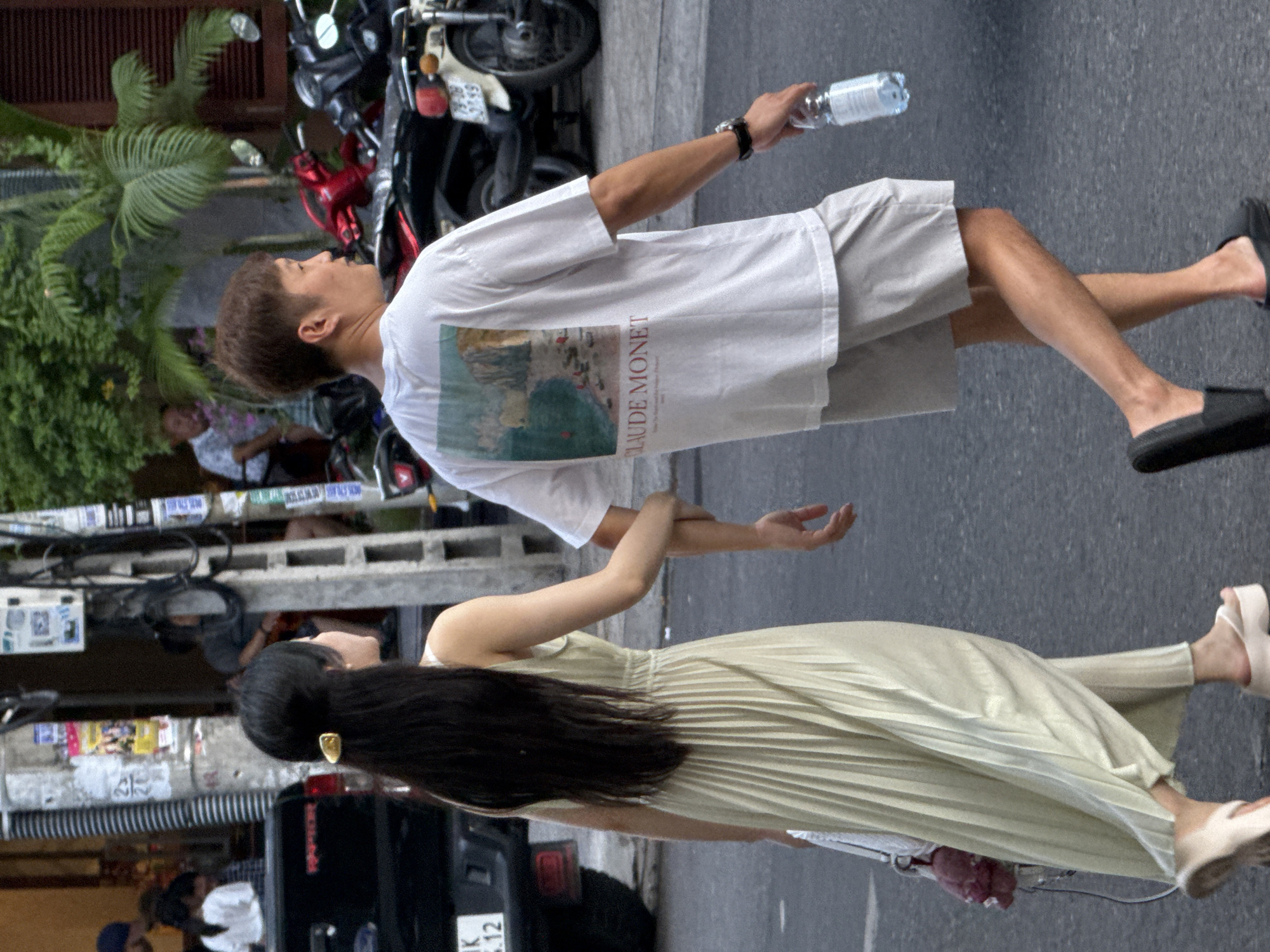 The image depicts a street scene in Nha Trang Ward, Vietnam, showing a young couple walking hand-in-hand. The woman, on the left, has long black hair and wears a flowing, light yellow pleated dress and platform sandals. She carries a white bag with a pink fuzzy accessory. The man, on the right, has short brown hair and is dressed in a white t-shirt featuring a landscape print by Claude Monet, and light gray shorts. He wears black slide sandals and carries a bottle of water in his right hand. They are walking on an asphalt road. In the background, there are motorcycles parked, a dark-colored vehicle, a utility pole with various posters and wires, and hints of buildings and foliage.