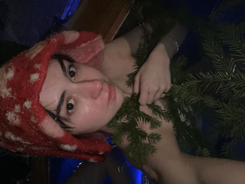 The image depicts a young adult female, partially submerged in dark blue water, likely an outdoor bath or hot tub. She has dark hair, brown eyes, and a flushed, wet face. She wears a red and white knitted or felted hat, patterned to resemble a mushroom cap, with small white particles (possibly snow or ice) visible on the hat and her hair. Coniferous branches, appearing to be fir or spruce, are draped across her chest and arms; she holds some of these branches with her right hand. A thin, light-colored bracelet is visible on her left wrist. The background is dark, suggesting a nighttime or low-light setting. The scene is located in Базарово, Russia.