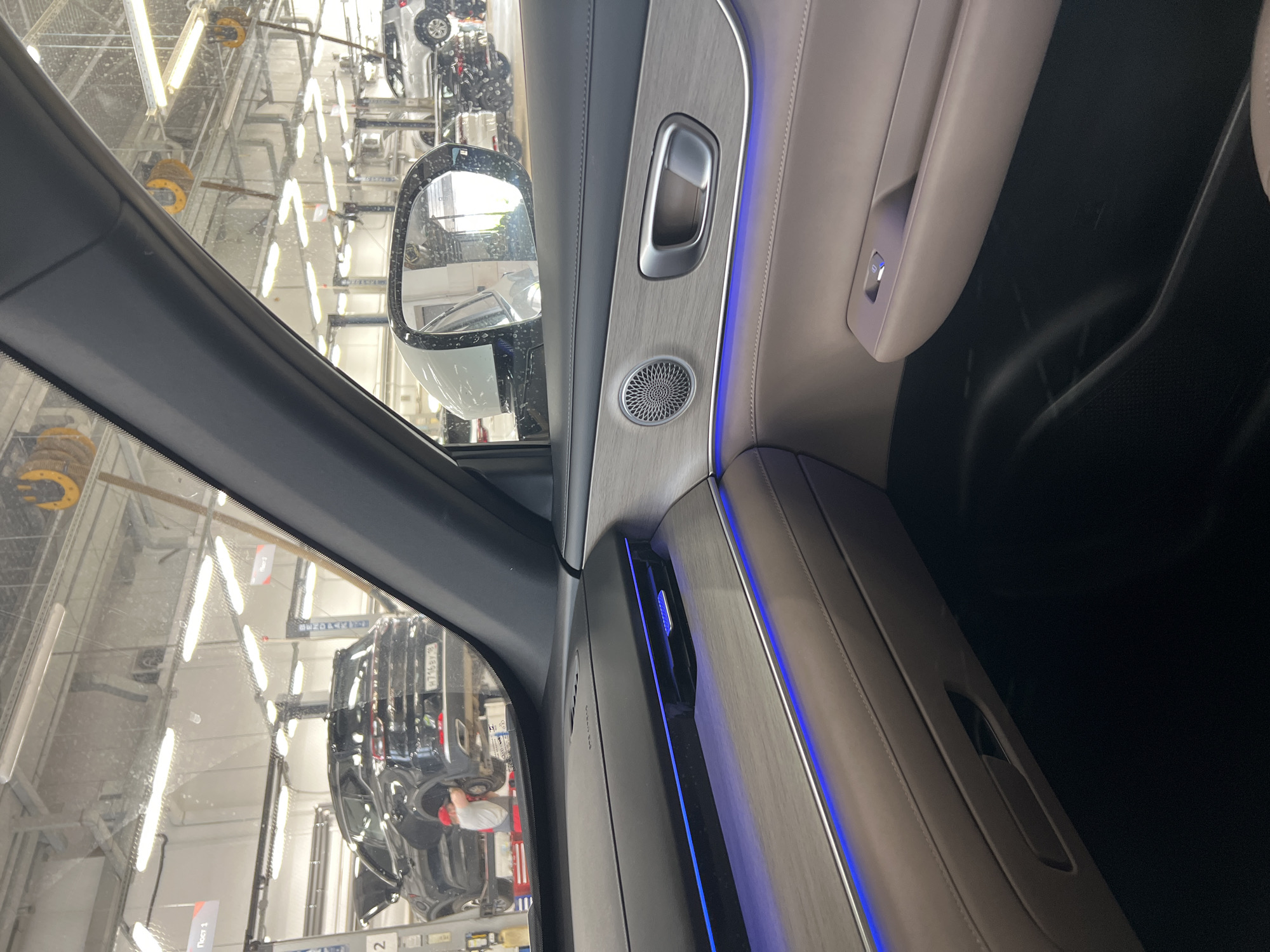 The photograph, taken from inside a modern passenger vehicle, depicts an auto service center in Izhevsk, Udmurt Republic, Russia. The location is confirmed by the license plate "М716ВУ 18" visible on a black SUV in the background, where "18" is the region code for the Udmurt Republic, and by Cyrillic text on signs such as "Пост 1" (Post 1), "Пост 3" (Post 3), and "БЕЛОРУС" (Belarus, possibly a brand or company name).
The interior of the vehicle from which the photo is taken features a beige and dark gray color palette with blue ambient lighting strips along a textured wood-grain trim piece on the dashboard and door panel. A circular speaker grille with a metallic mesh pattern, a metallic door handle, and an air vent are visible on the passenger side. A window control button with a blue indicator light is situated on the lower part of the door panel. The vehicle's side mirror reflects a portion of the service center, including a white SUV on another lift. The windshield shows visible dust or splatter.
Beyond the vehicle's interior, the auto service center is brightly illuminated by numerous overhead fluorescent light fixtures. A black SUV, resembling a Chery Tiggo 8 Pro, is elevated on a hydraulic lift, with its hood open. A lone individual, wearing a red cap and a white t-shirt, is engaged in servicing the rear wheel area of this black SUV. Multiple other hydraulic vehicle lifts are discernible throughout the facility, one of which holds a white SUV in the background. Yellow industrial hose or cable reels are suspended from the ceiling. The walls of the service bay are painted white.