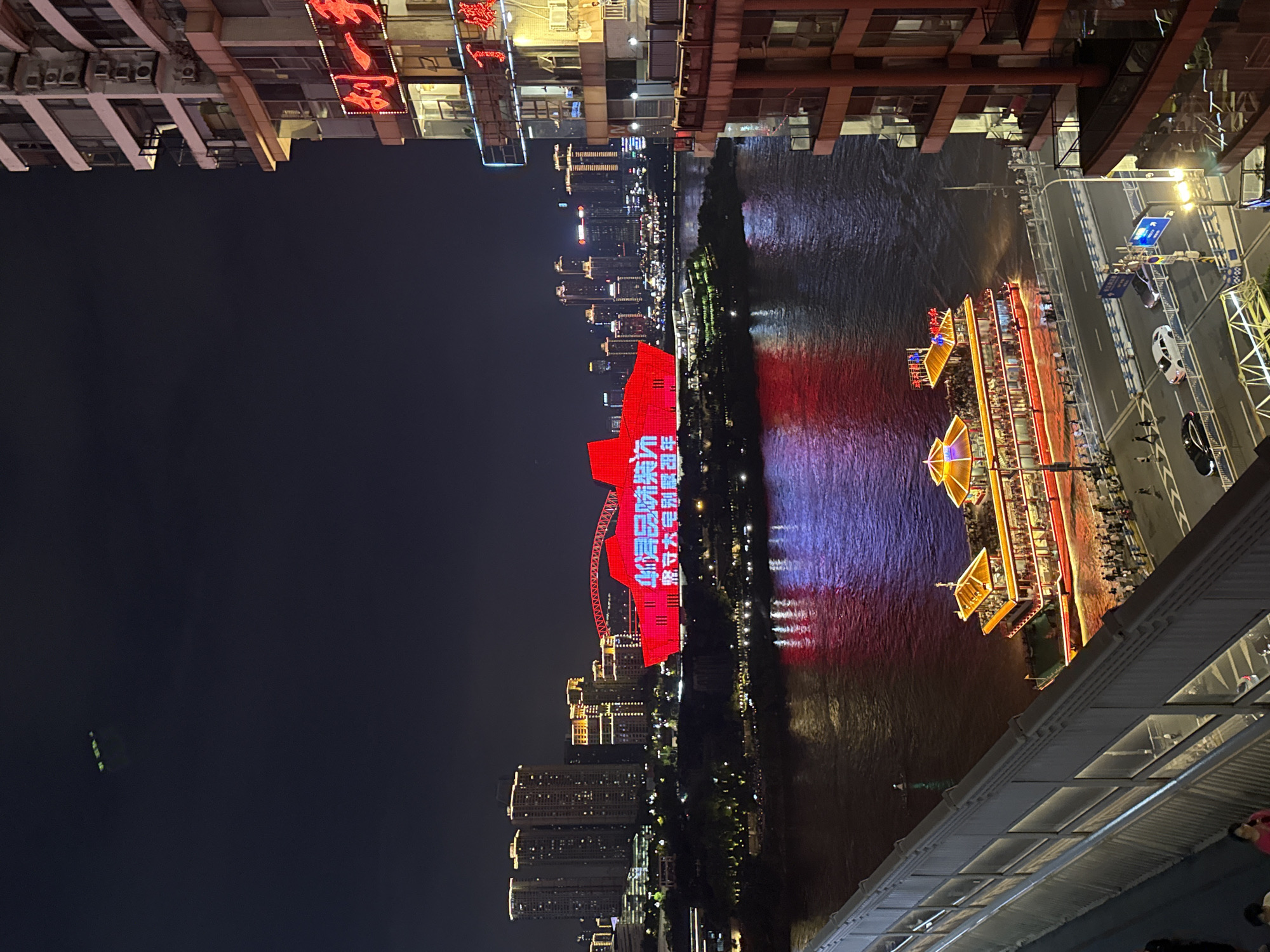 The image depicts a nighttime view of the Yuzhong District in Chongqing, China, focusing on the Yangtze River. On the river, a multi-decked, illuminated tourist boat with traditional Chinese architectural elements is docked, its lights casting a warm glow on the water. The river's surface reflects the vibrant red and purple lights from the illuminated buildings and structures along the riverbank and in the distance.
Dominating the skyline across the river is a striking red, illuminated building with Chinese characters displayed prominently, likely a landmark or advertisement. Several modern high-rise buildings, illuminated with warm yellow and white lights, form the city's backdrop. In the foreground, on the right side of the image, a tall, modern building with balconies and numerous windows is visible, its illuminated facade contributing to the overall nighttime cityscape.
A street runs along the riverfront, with several cars and a few pedestrians visible. The walkway beside the river appears to be lined with barriers and lights, and a bridge structure is partially visible in the lower left of the frame. The sky is dark, indicative of nighttime, with a faint green light source observed in the upper left quadrant, possibly an aircraft or drone. The scene is characterized by a strong interplay of artificial lights against the dark night sky and river.