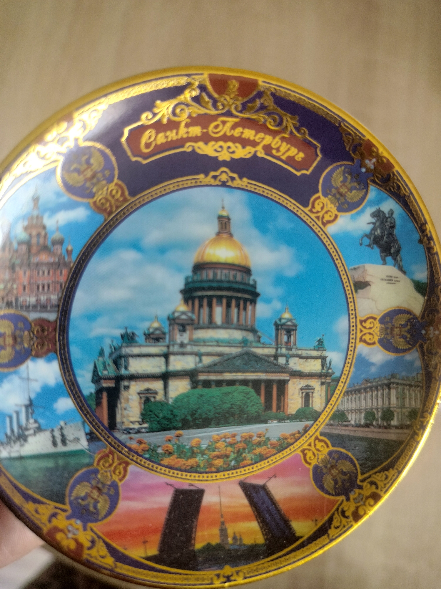 The image shows a decorative plate featuring a montage of St. Petersburg landmarks. The plate has a gold rim and a dark blue border decorated with gold scrollwork and imperial eagles. In the center, a circular scene depicts St. Isaac's Cathedral with its golden dome, surrounded by other architectural elements. The outer edge of the plate shows various vignettes: a pink and orange sunset with a raised bridge and the Peter and Paul Fortress spire, a large building resembling the Winter Palace, a ship, and a statue of a man on horseback, likely the Bronze Horseman. The plate is not located in Yashkino, Russia; it depicts scenes from St. Petersburg. There are no people visible in the image.