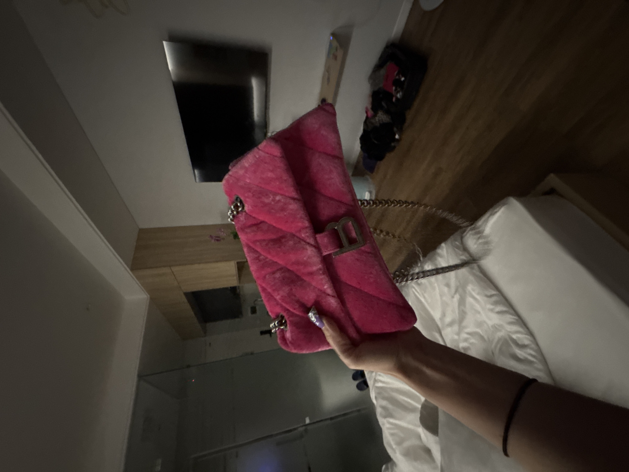 A person's arm and hand are holding a bright pink quilted handbag with a silver chain strap and a silver buckle. The person's fingernails are painted purple and decorated with glitter. The handbag is held in front of a white duvet on a bed. In the background, a large black flat-screen television is mounted on a grey wall above a wooden shelf. To the right of the television, there is a wooden closet or cabinet. On the wooden floor in the lower right corner, there is a pile of clothing and a black duffel bag. A glass-paneled door is visible on the left side of the image, suggesting the scene may be inside a hotel room or apartment. The lighting is dim, casting shadows.