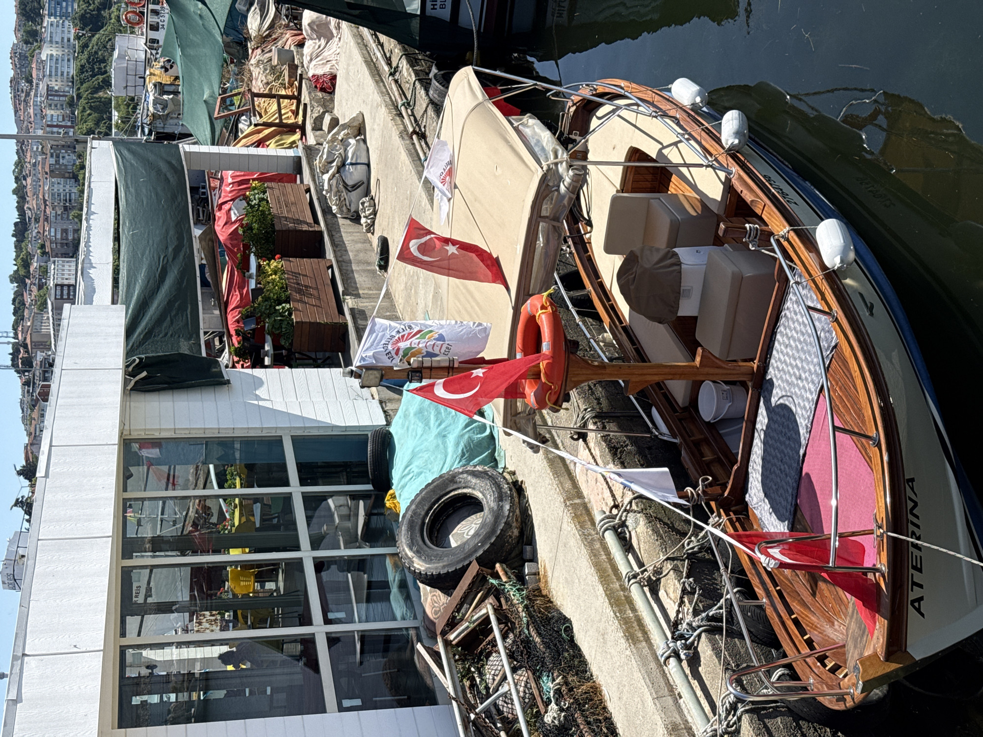 This image shows a harbor scene in Şile, Turkey, on a sunny day. A wooden boat named "ATERINA" is moored at a dock, facing towards the viewer. The boat has a tan canopy, seating, and several flags, including the Turkish flag. The dock area is cluttered with various objects, including fishing nets, a large tire, and tarps covering materials. In the background, a white building with large windows is visible, and beyond that, a hillside covered with buildings and trees. The water is dark and calm, reflecting the sky and surrounding structures. The overall impression is of a working harbor with boats and maritime equipment.