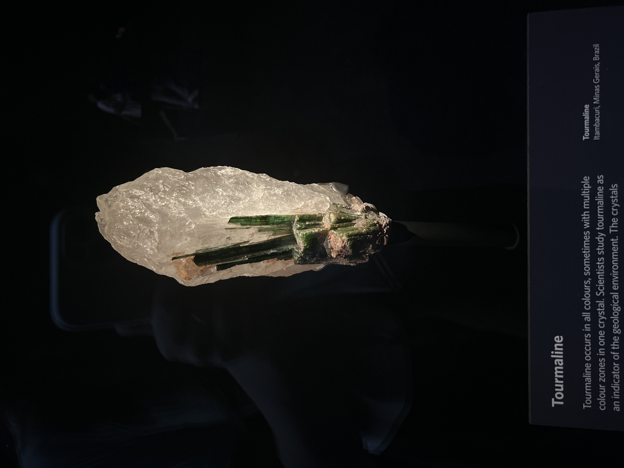 A museum exhibit displays a large, rough, translucent white crystal with several dark green tourmaline crystals embedded within it. The exhibit is dimly lit, with the crystal illuminated from the front, casting a shadow. In the background, two blurry figures are visible, suggesting this is a public space like a museum. A dark blue plaque at the bottom of the image contains text related to the display. The location of this exhibit is not determinable from the image alone, and therefore the city of London, United Kingdom, cannot be specified.