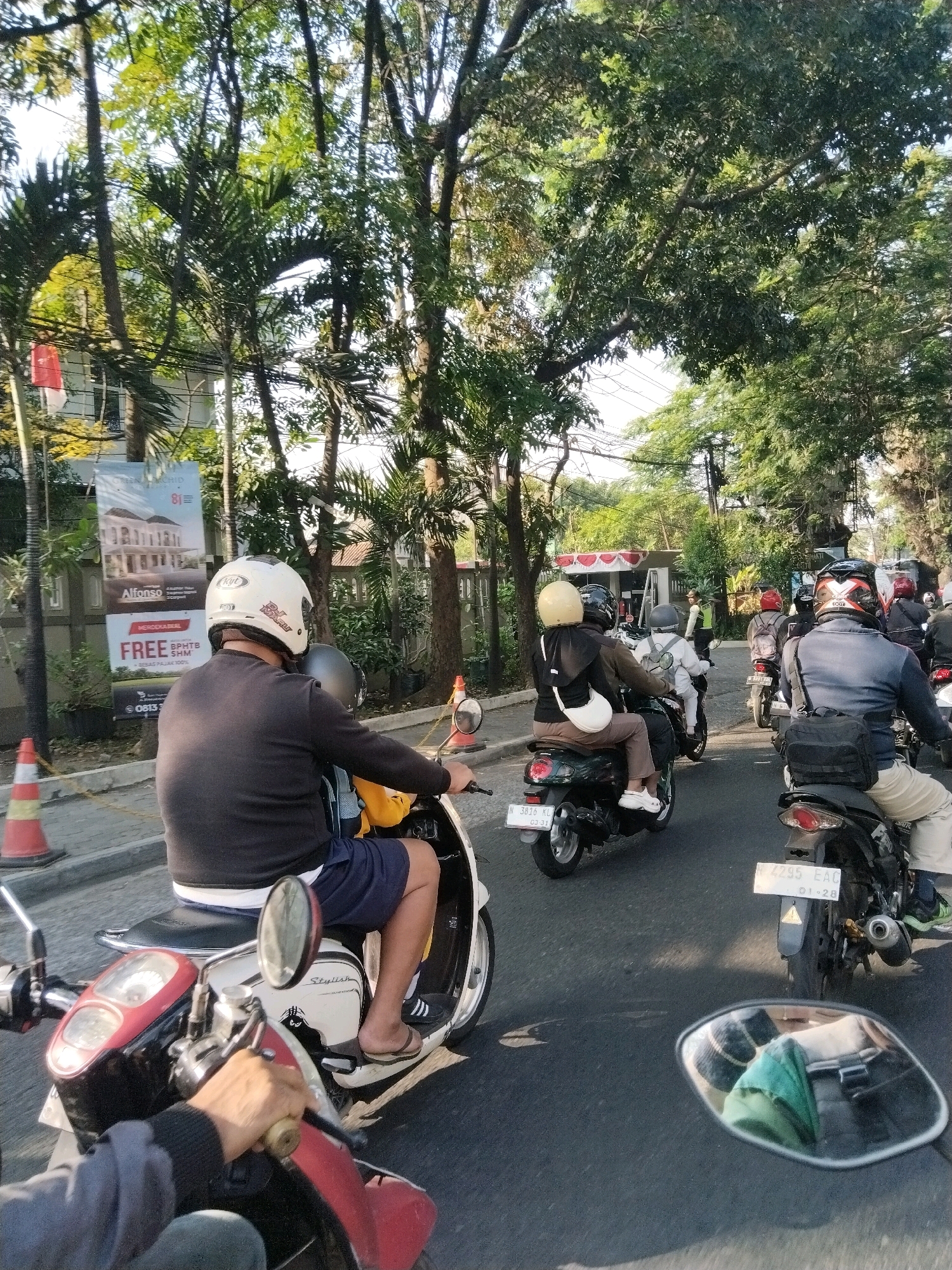 The image depicts a street scene in Banjararum, Indonesia, characterized by moderate traffic. In the foreground, a person on a scooter, wearing a white helmet and a dark long-sleeved shirt with blue shorts, is visible from the back. Their right hand is on the scooter's handlebar. The scooter itself is white and red. To the left, another scooter's handlebar and part of the rider's arm are visible. In the middle ground, several other scooters with riders are moving in the same direction. One rider wears a yellow helmet, another a dark helmet. Behind them, more scooters and riders are seen, with some wearing helmets and backpacks. The environment features lush trees lining the street, with some buildings and billboards visible in the background. The lighting suggests it is daytime, likely in the morning or afternoon, with sunlight filtering through the foliage. Traffic cones are present on the side of the road, indicating potential road work or traffic management. The overall atmosphere is that of a typical urban commute in Indonesia.