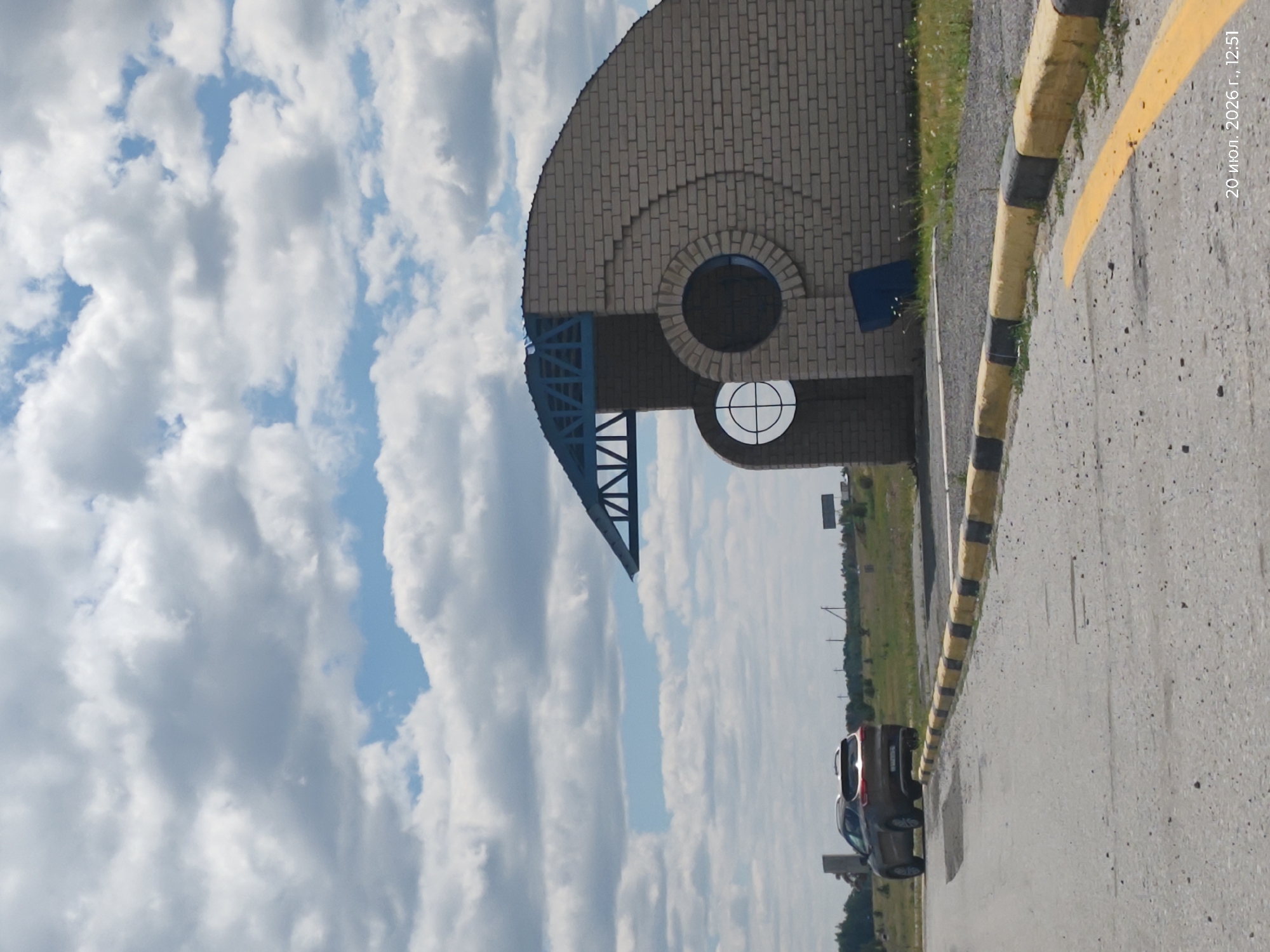This is a photograph taken on July 20, 2026, at 12:51 PM, in Aidarly, Kazakhstan. The scene depicts an exterior view of a building constructed with light-colored bricks. The building has a unique curved design and features two circular windows. A blue, angular roof structure is attached to the building. In the foreground, a road or paved area is visible, bordered by a curb with alternating yellow and black markings. A dark-colored SUV is parked on the left side of the road, facing away from the viewer. The background shows a flat, grassy landscape with scattered trees and utility poles. The sky is predominantly filled with large, white, puffy clouds against a blue backdrop.