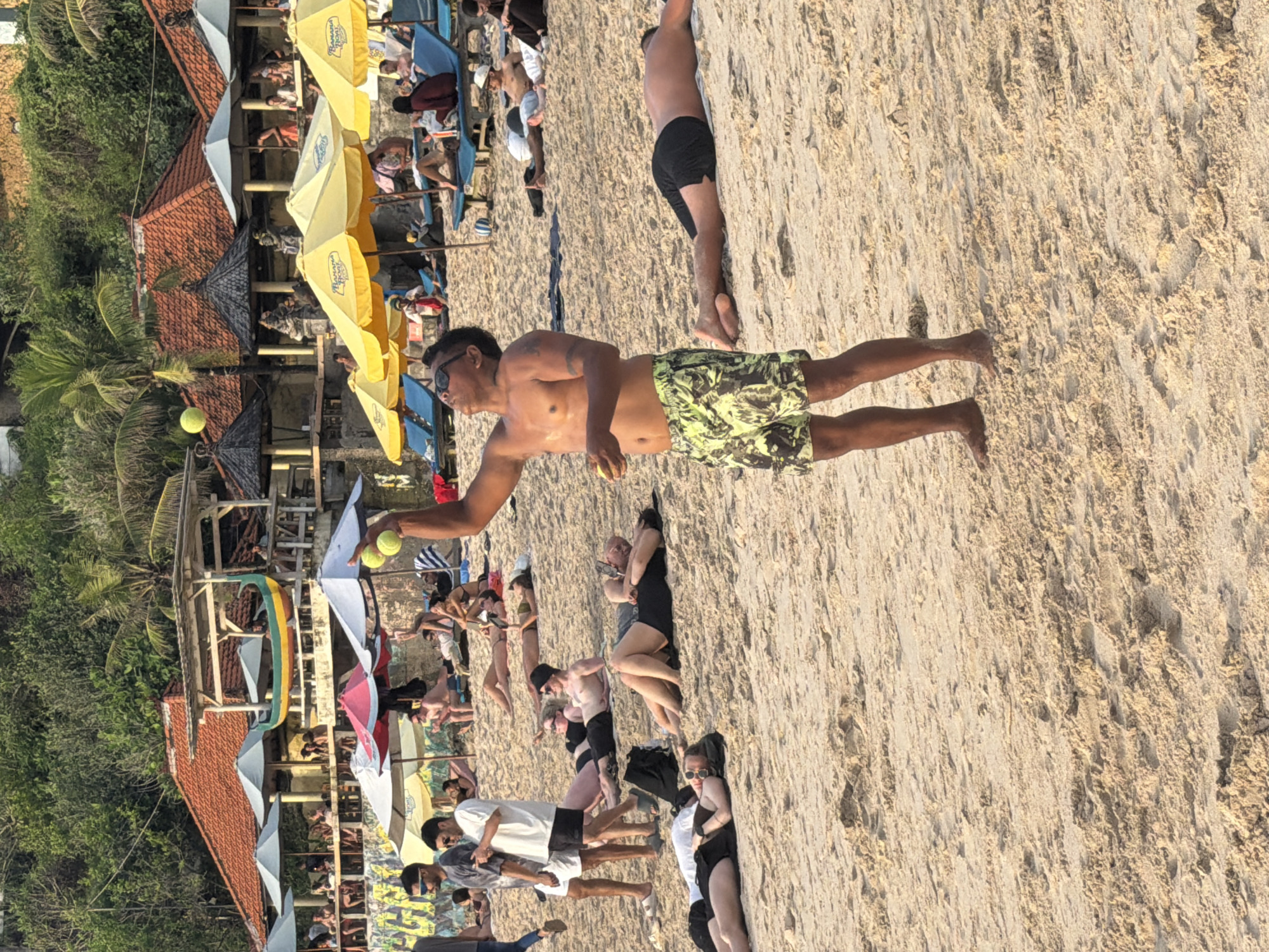 The image depicts a crowded beach in Kuta Selatan, Indonesia. The scene shows various individuals engaged in leisure activities. A shirtless man in patterned shorts is throwing a tennis ball. Several people are sunbathing or walking along the sandy beach. Rows of beach umbrellas provide shade for beachgoers. In the background, there are buildings with red tile roofs and green trees, suggesting a coastal environment. The overall atmosphere is vibrant and summery.