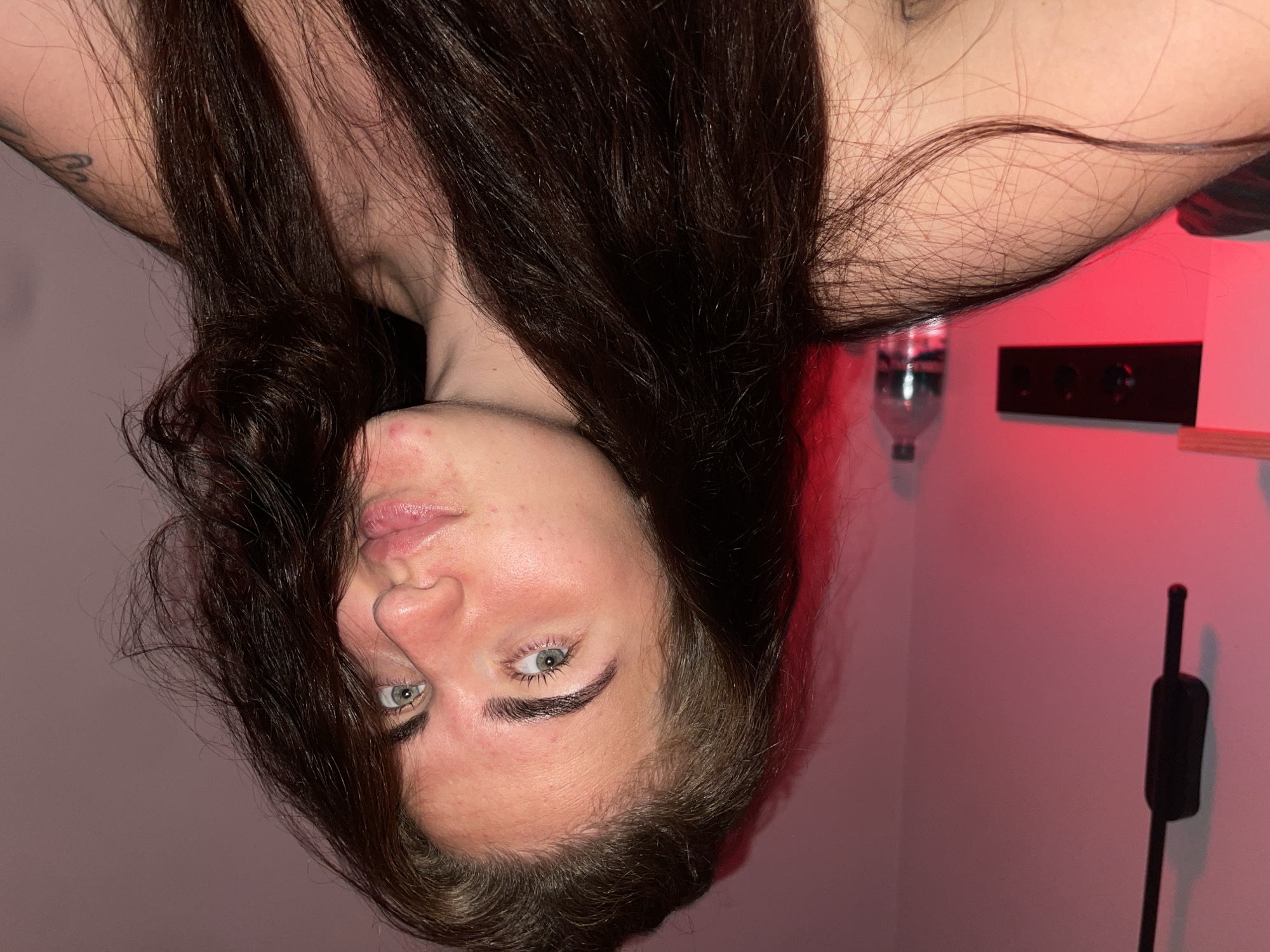 In the city of Мосметаллоконструкция Онт, Russia, a young woman with long, dark brown hair is captured in a close-up selfie. She is indoors, likely in a bedroom, as indicated by the soft, pinkish lighting that bathes the scene. The background features a pale pink wall, a black electrical device hanging on the wall, a shelf with a black sound system, and a plastic water bottle on the shelf. The woman's face is turned slightly towards the camera, with her light green eyes looking directly into the lens. Her facial features include light skin with some redness and minor blemishes. Her lips are slightly parted, giving her a playful expression. Her dark hair is mostly spread around her shoulders and chest, with some strands falling across her face. The overall impression is a casual, intimate moment captured in a domestic setting.