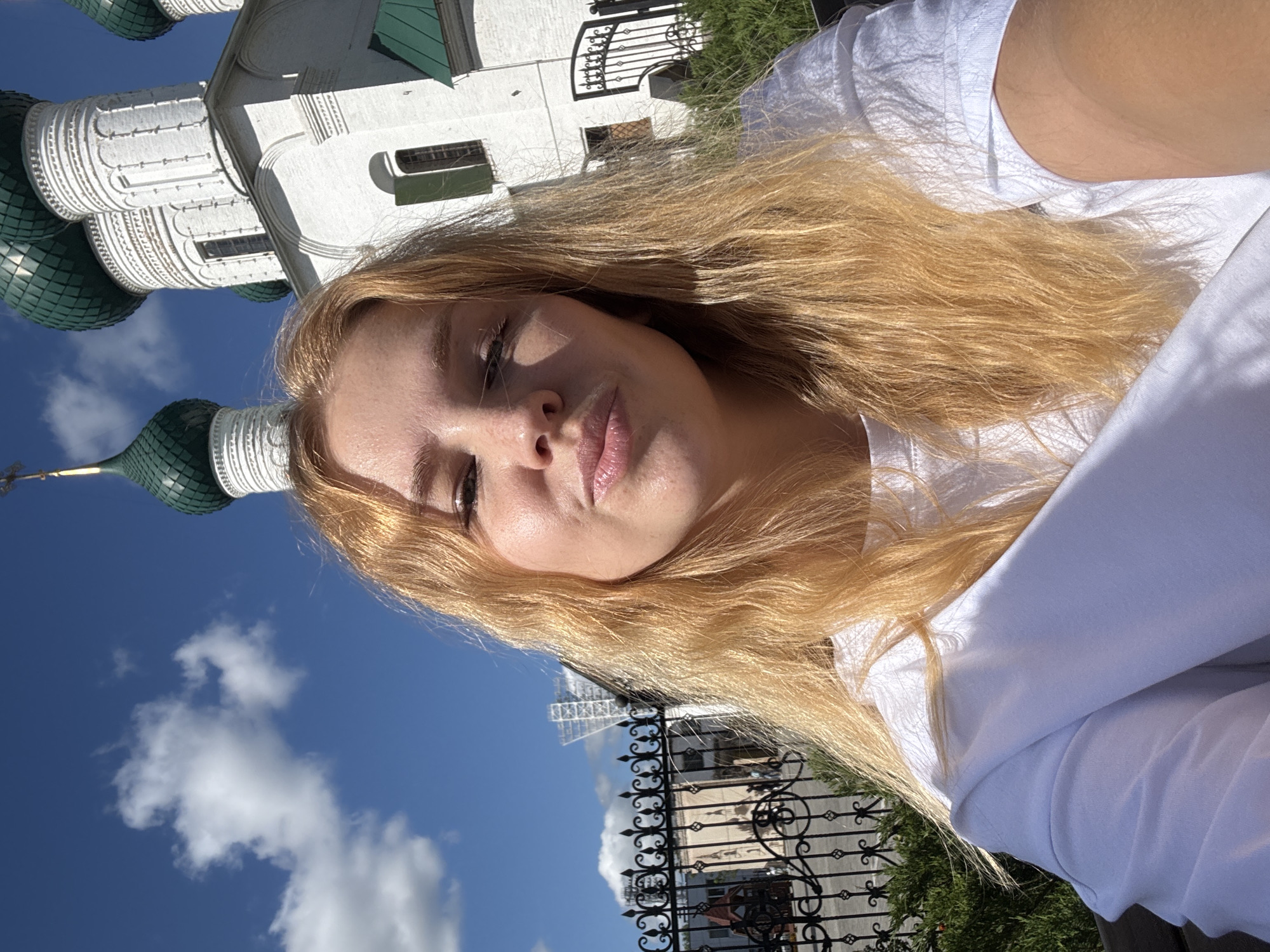 In Zhukovo, Russia, a young woman with blonde hair, wearing a white t-shirt, takes a selfie in front of a white church with green domes. The sky is blue with some clouds, and a black wrought-iron fence is visible in the foreground. The woman's face is close to the camera, and she is making a duck face. The church appears to be an Orthodox Christian church, judging by its architectural style. The overall scene suggests a sunny day with a pleasant atmosphere.