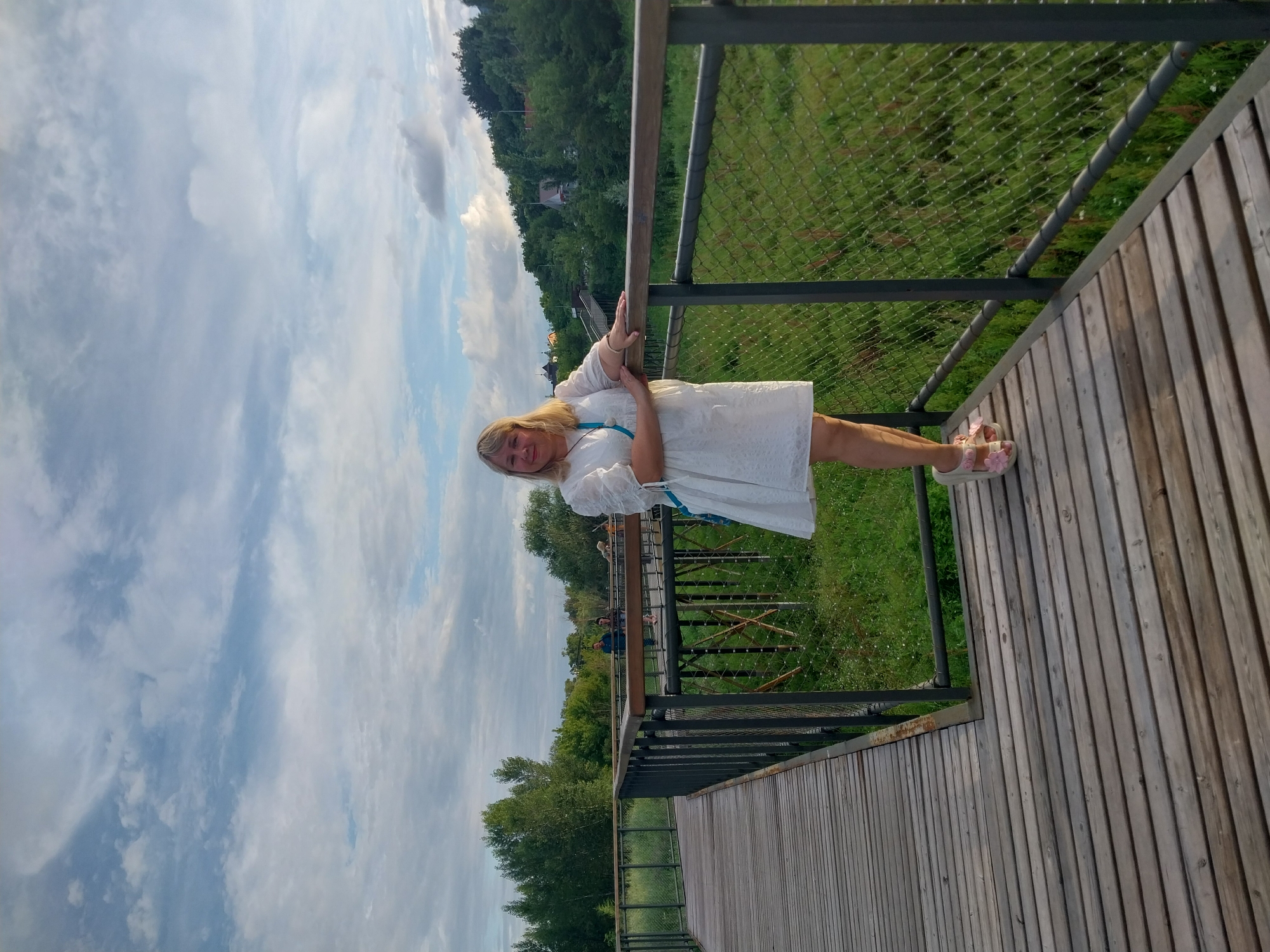 A blonde woman wearing a white dress and pink sandals stands on a wooden bridge, leaning on the railing. The bridge is elevated above a field of green grass and reeds, with trees and a hill in the background. The sky is cloudy, with patches of blue. In the distance on the bridge, two people are walking away from the camera. The scene appears to be in a park or nature reserve, possibly in Orenburg, Russia, given the flora and landscape.