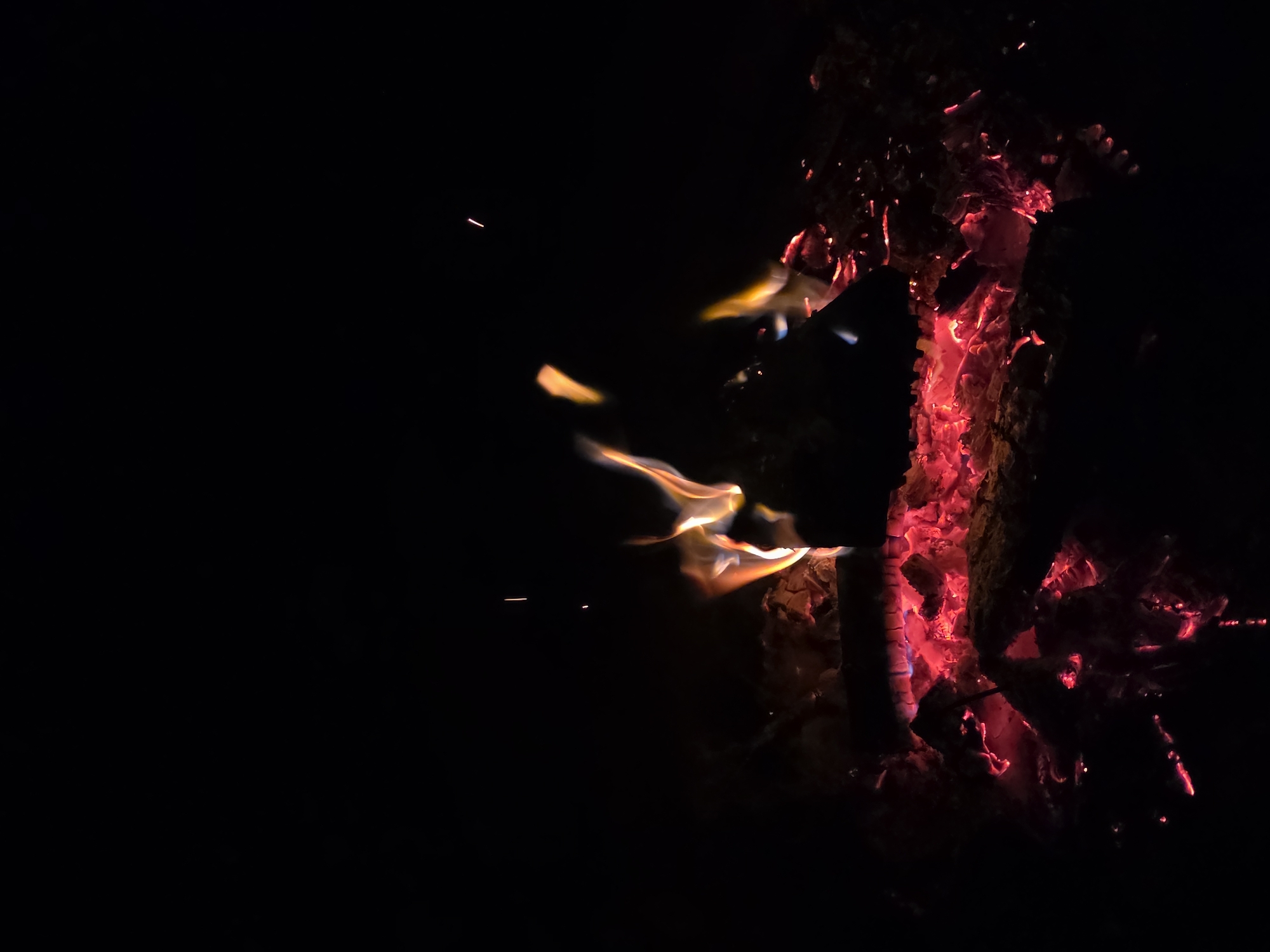 The image shows a close-up view of a campfire at night. The flames are primarily orange and yellow, with some white highlights, licking upwards from glowing red embers and charred wood. Sparks are visible ascending into the dark background. The scene appears to be set in an outdoor environment, possibly a campsite or backyard, given the presence of a fire pit and logs. The darkness of the background suggests it is nighttime, enhancing the glow of the fire. There is no indication of people or any specific location within the city of Строитель-нефтяник Снт, Russia.