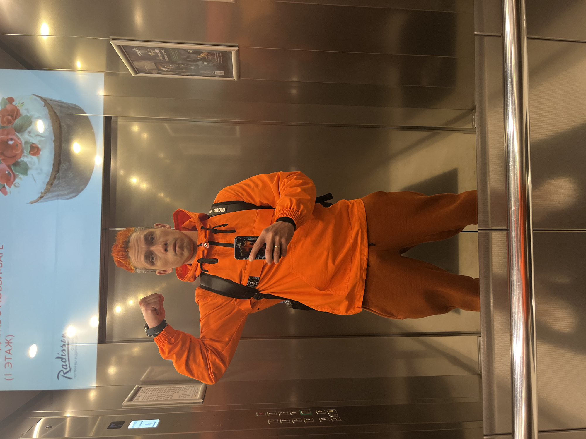 A man is standing in an elevator. He has orange hair and is wearing an orange jacket and pants. He is holding a phone in his right hand and flexing his left arm. The elevator has a Radisson poster and buttons for the floors. The elevator is likely located in the city of Moscow, Russia.