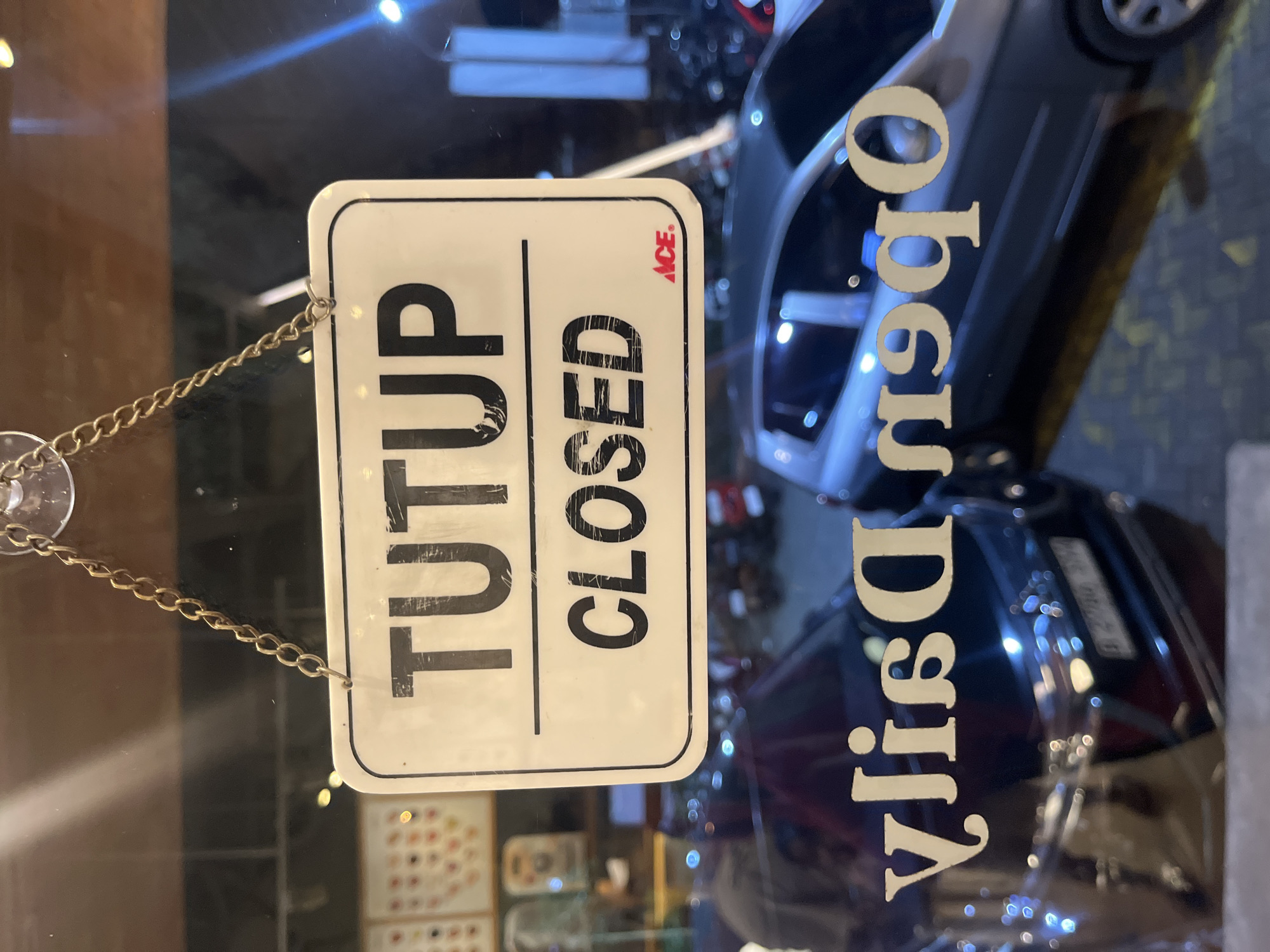 The image shows a "CLOSED" sign hanging on a glass door. The sign has "TUTUP" (Indonesian for closed) on the left side and "CLOSED" on the right side with "ACE" logo on the upper right side. It is suspended by a gold chain. Reflections on the glass door reveal that it is a store named "Djay's". The store's location is likely in South Tangerang, Indonesia, given the Indonesian translation of the word "closed". The details within the store are difficult to make out.
