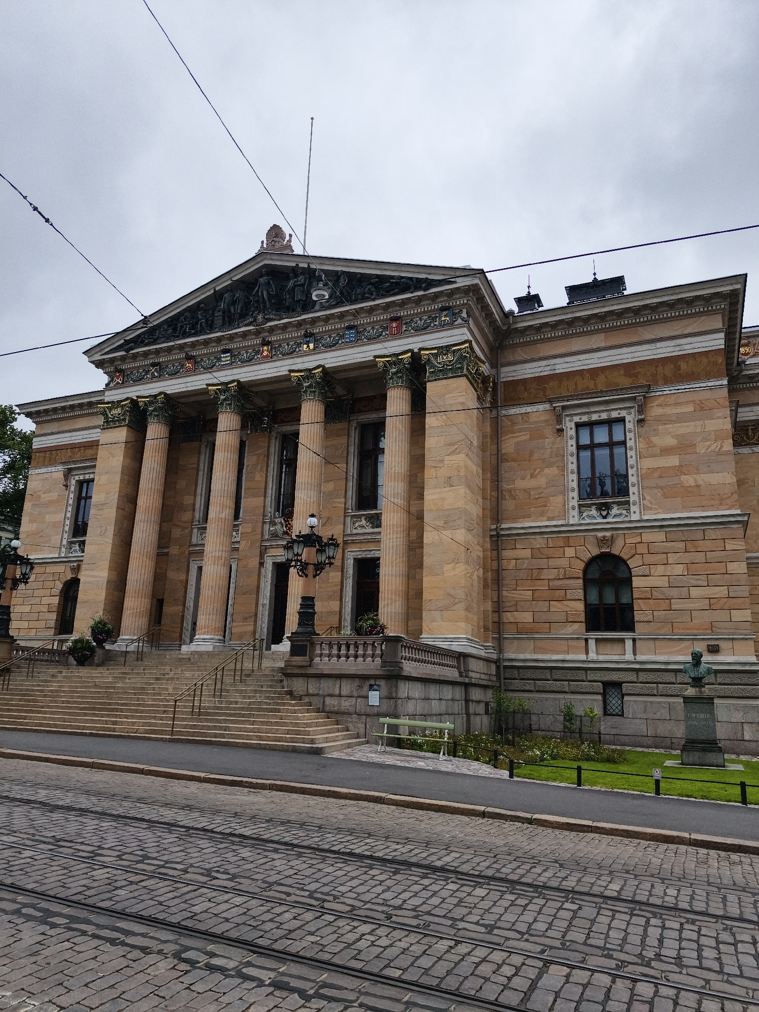 The image shows the Helsinki Central Library Oodi, a public library in Helsinki, Finland. The building features neoclassical architecture with large columns, ornate details, and a mansard roof. In the foreground, tram tracks are visible on a cobblestone street, indicating an urban environment. A few people are standing near the entrance, and some potted plants are placed on the steps leading up to the building. The sky is overcast, suggesting a cloudy day.