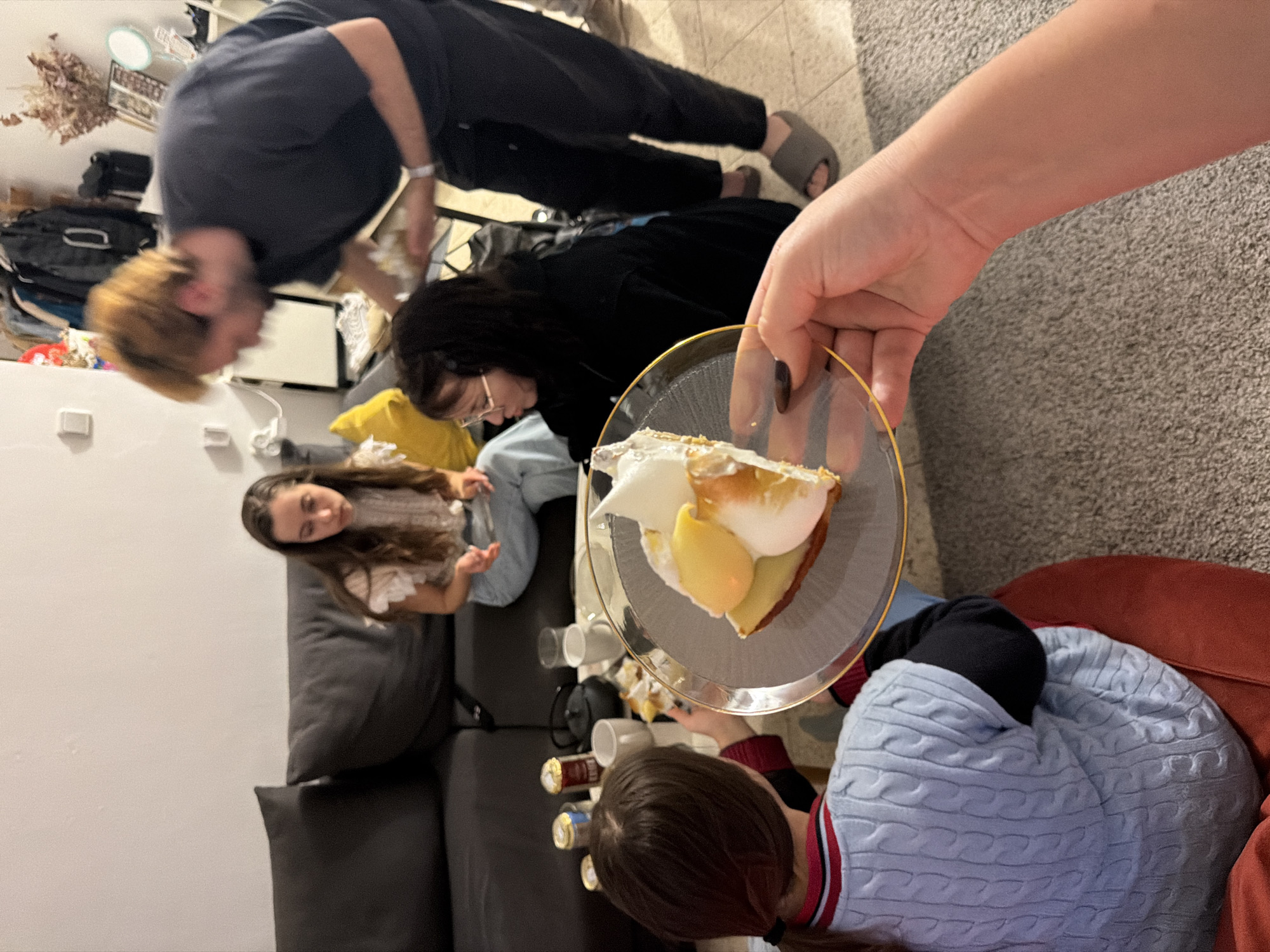 A gathering indoors shows several people. One person holds a slice of lemon meringue pie on a glass plate with a gold rim. Several empty mugs and beverage cans sit nearby on a dark-colored sofa. The setting includes people sitting on couches and chairs, indicating a casual social occasion. A partial glimpse of Ramat Gan, Israel, is indicated in the metadata.