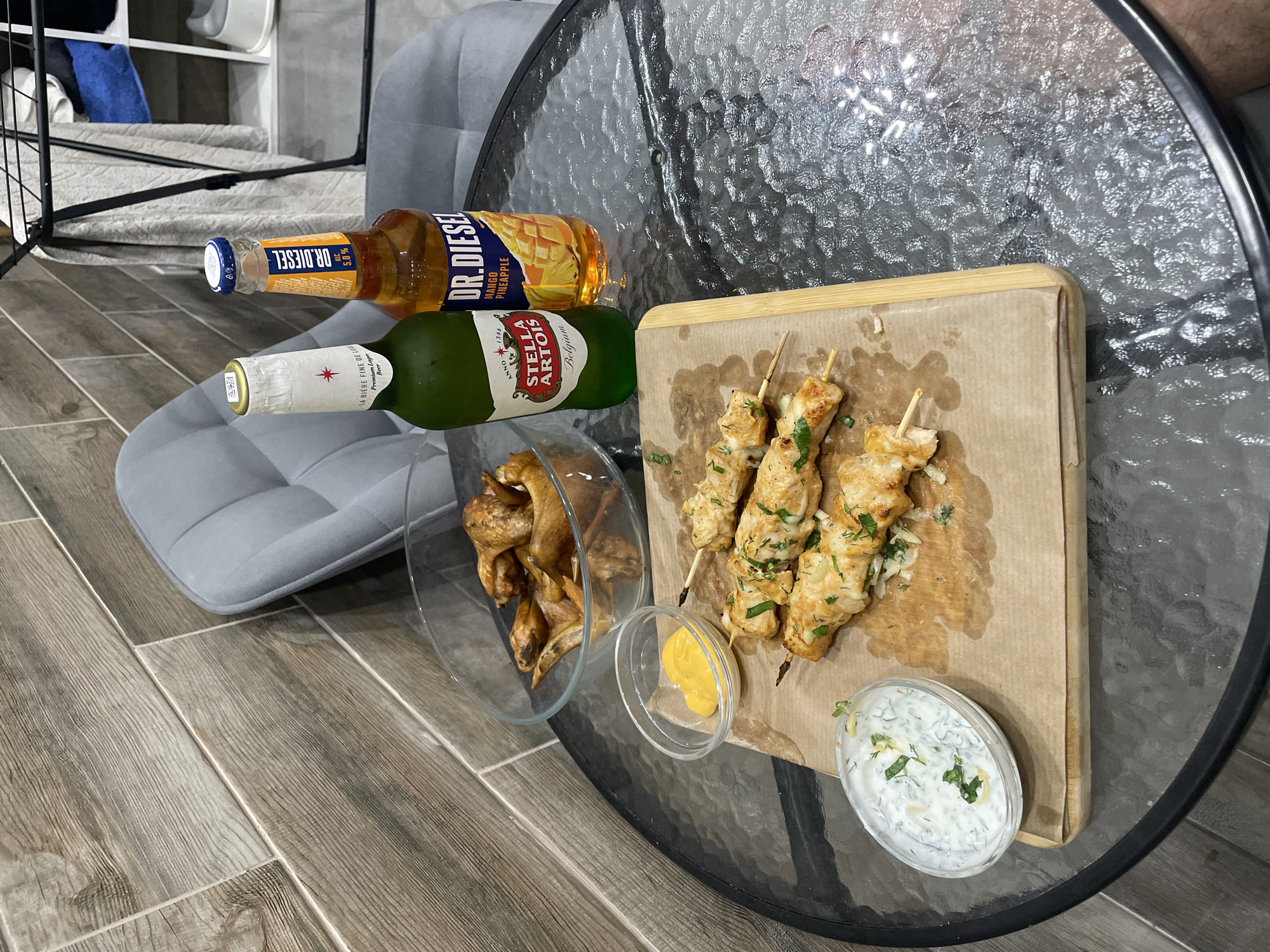 The image shows a glass table with food and drinks. There are chicken skewers on a wooden board, with two small bowls of sauces, one yellow and one white. There is also a bowl of what appears to be chicken wings. Two bottles of alcoholic beverages are present: one is a green bottle of Stella Artois beer, and the other is a bottle of Dr. Diesel Mango Pineapple drink. The table is next to a grey chair on a wooden floor. The photograph appears to be taken indoors. It is impossible to tell if it is Эстачи, Russia from the image.