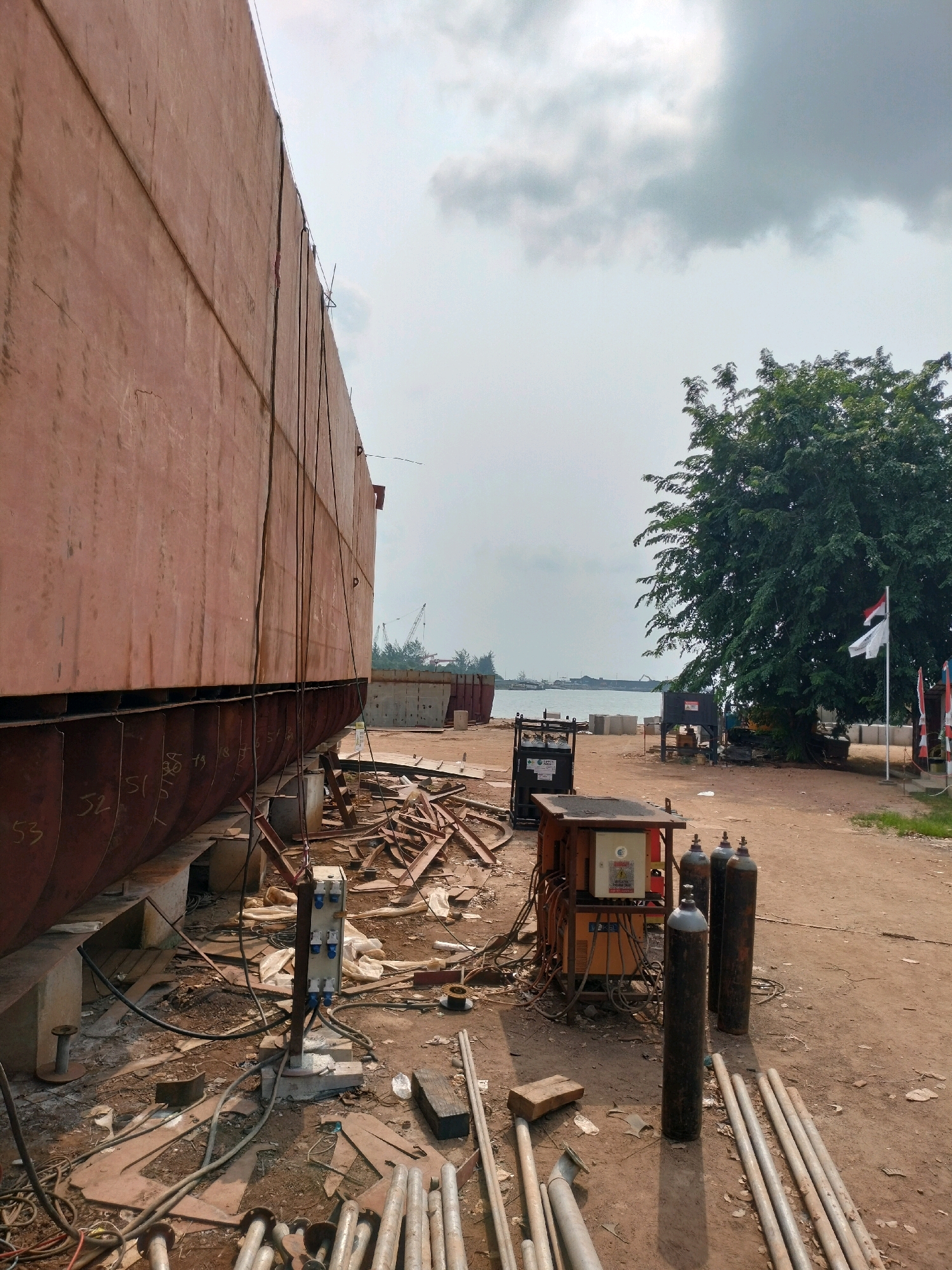 A dry dock in Batam, Indonesia, shows the immense hull of a ship under construction or repair. The ship's hull, made of large, rusty-red metal plates, dominates the left side of the frame, supported by robust grey blocks. The lower portion of the hull is marked with white numbers, including "53," "52," and "51," indicating sections or identification.
The ground is a dusty, reddish-brown earth, littered with construction debris: scrap metal pieces, lengths of pipe, and tangled electrical cables. In the center-right of the image, a welding machine sits on a wheeled cart, connected to various electrical cords and hoses. Several tall, dark cylinders, likely compressed gas tanks (oxygen or acetylene), stand vertically near the welding equipment. A grey, industrial electrical junction box with multiple outlets is positioned on a metal pole closer to the ship.
In the background, a vast expanse of water stretches towards a hazy horizon, dotted with distant ships and cranes. A large, verdant tree with dense foliage stands prominently on the right, its branches reaching towards the overcast sky. Several flags, including one with red and white stripes, are visible near the tree. The sky is a muted grey, with scattered clouds suggesting a potentially humid or rainy atmosphere common in the region. The scene depicts an active but somewhat chaotic industrial environment focused on shipbuilding or maintenance.